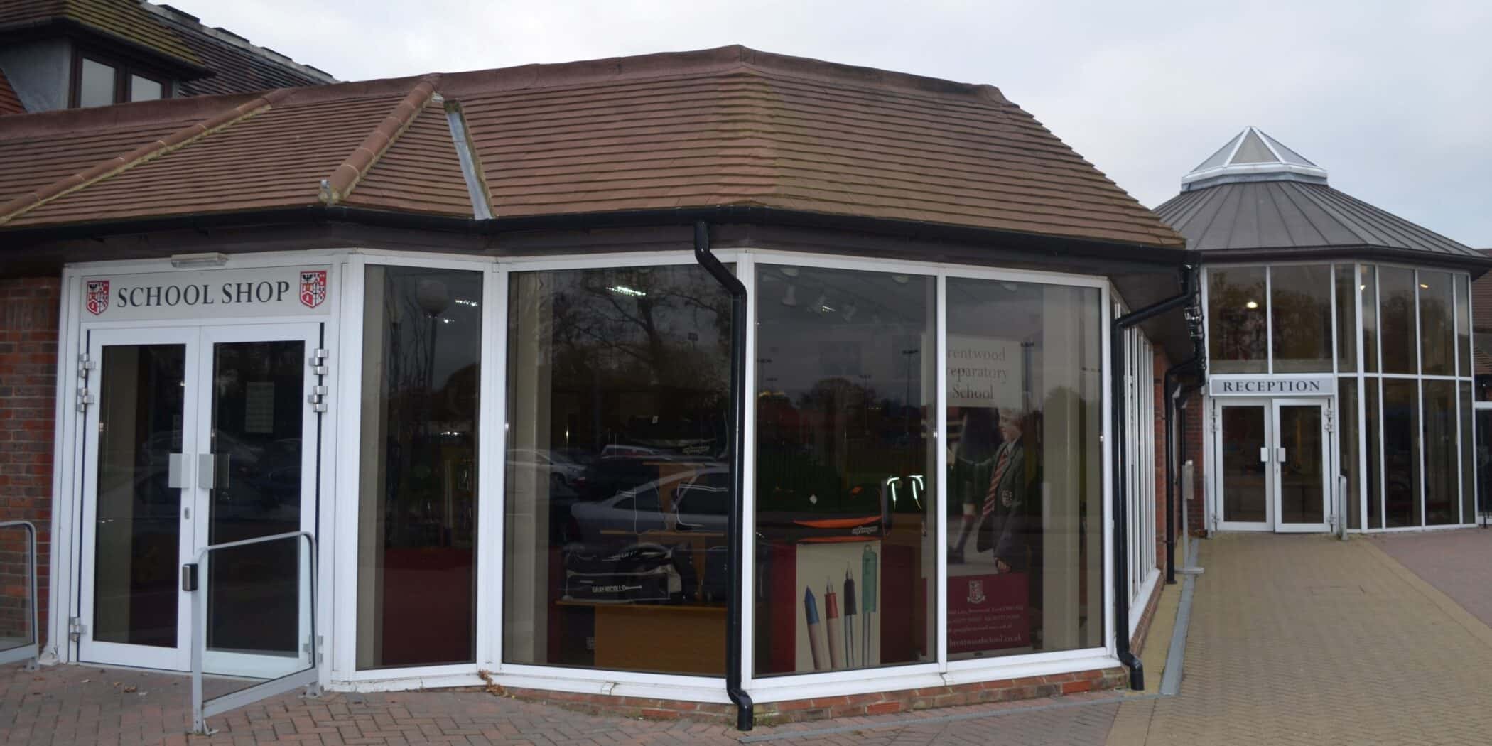 A modern brick building with large glass windows houses a school shop on the left, displaying uniforms and school items inside. To the right, a separate entrance is labelled Reception.