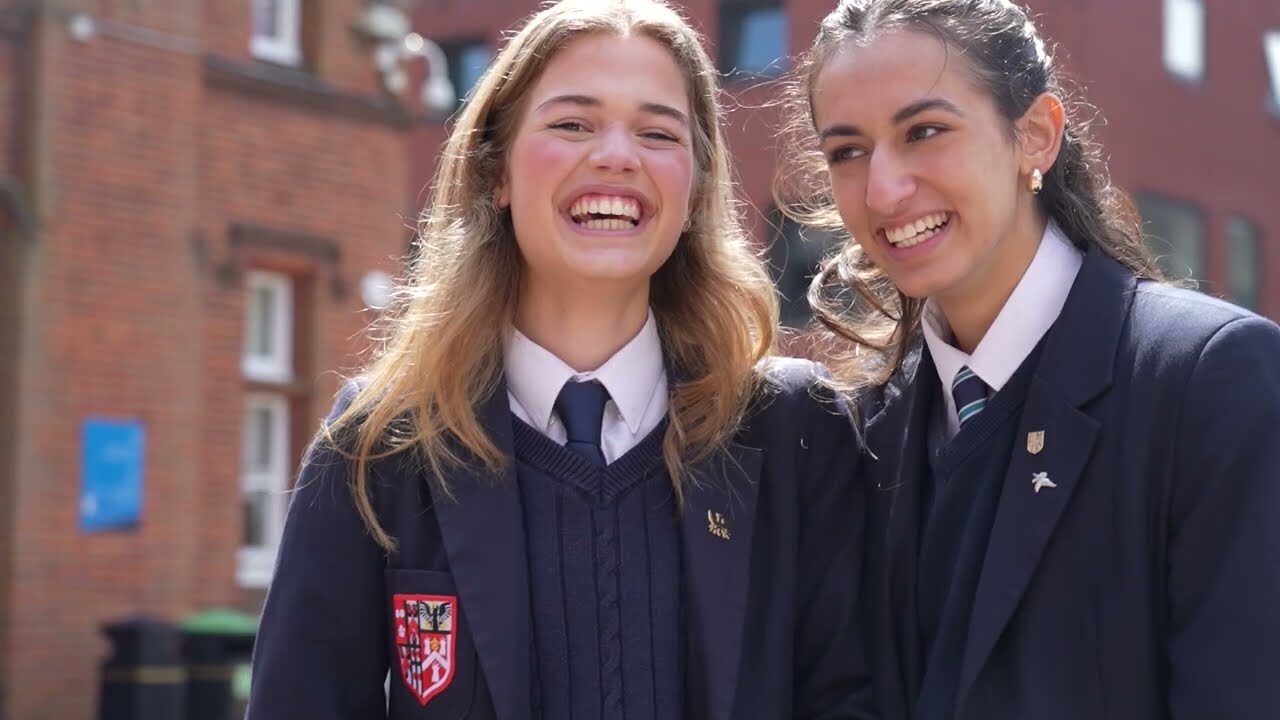 Two Brentwood School Sixth Form students in dark uniforms and ties stand close together outside a brick building, smiling and laughing.