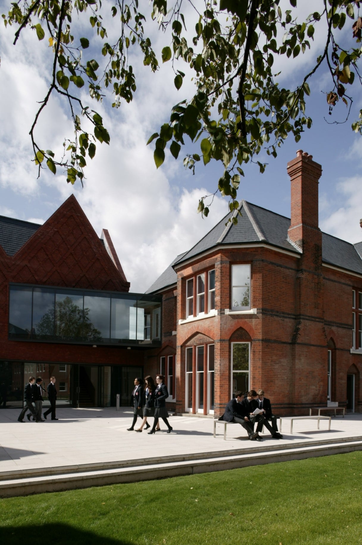 Students in uniforms walk and sit on benches outside a modern red-brick school building with large windows, on a sunny day with green grass and leafy branches in the foreground.