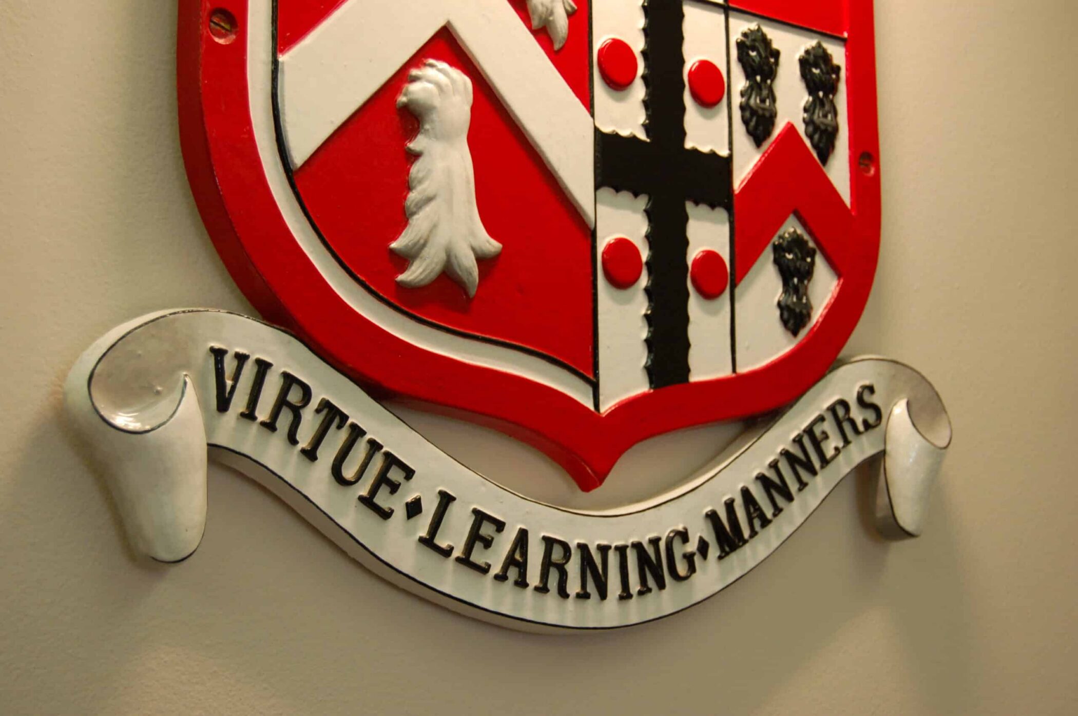 A close-up of a red and white shield-shaped crest with a banner underneath that reads VIRTUE • LEARNING • MANNERS in bold black letters.