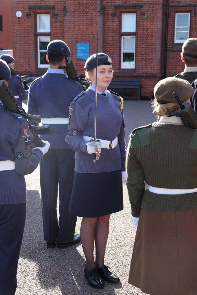 A uniformed cadet stands holding a sword during an outdoor Sixth Form Co-Curricular and Enrichment inspection, facing another cadet. Several others are in formation, with a brick building in the background.
