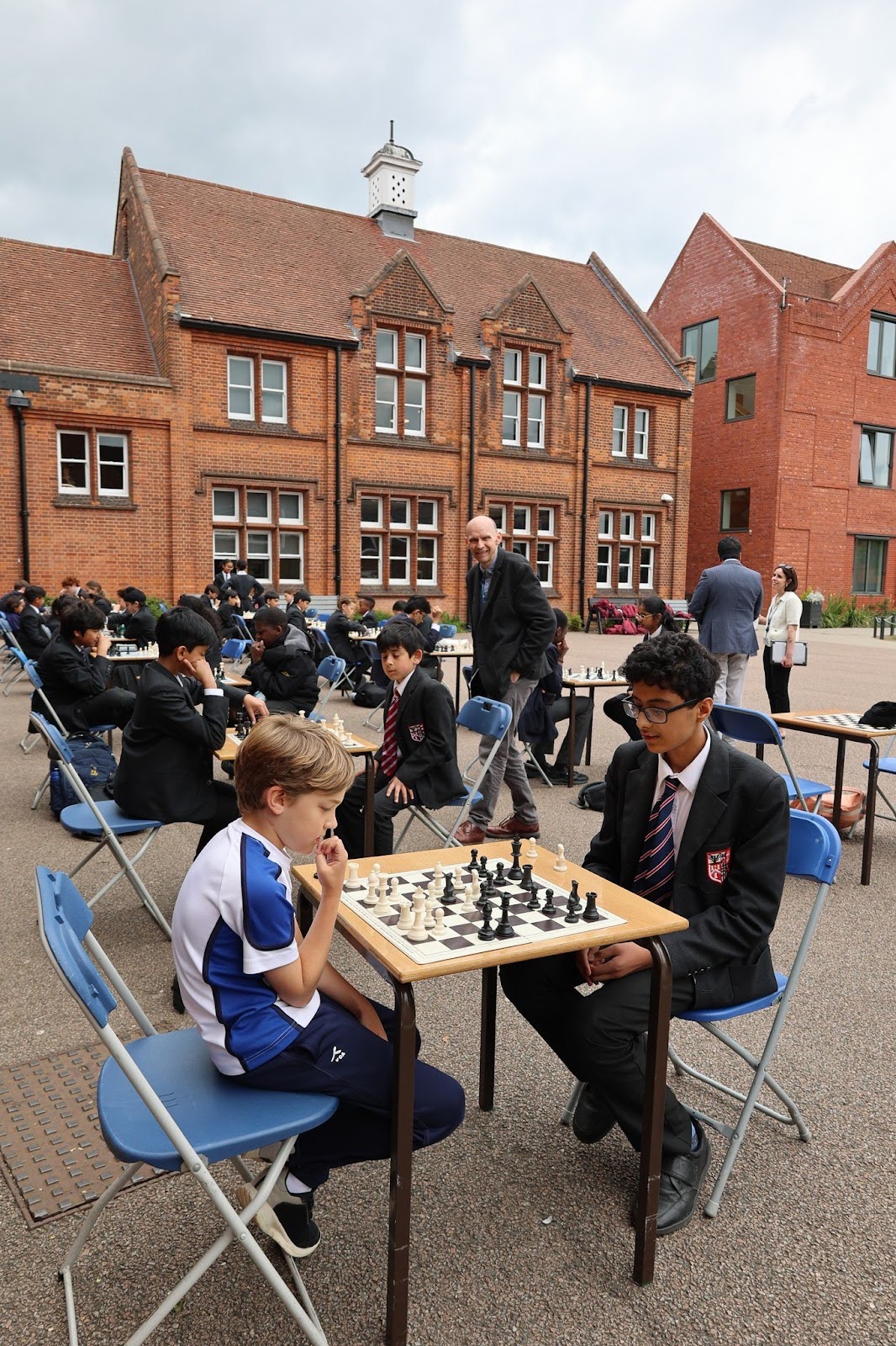 Students play chess on folding tables and chairs outdoors in a school courtyard, with a brick building in the background. Other students and adults observe or take part in the event under a cloudy sky.