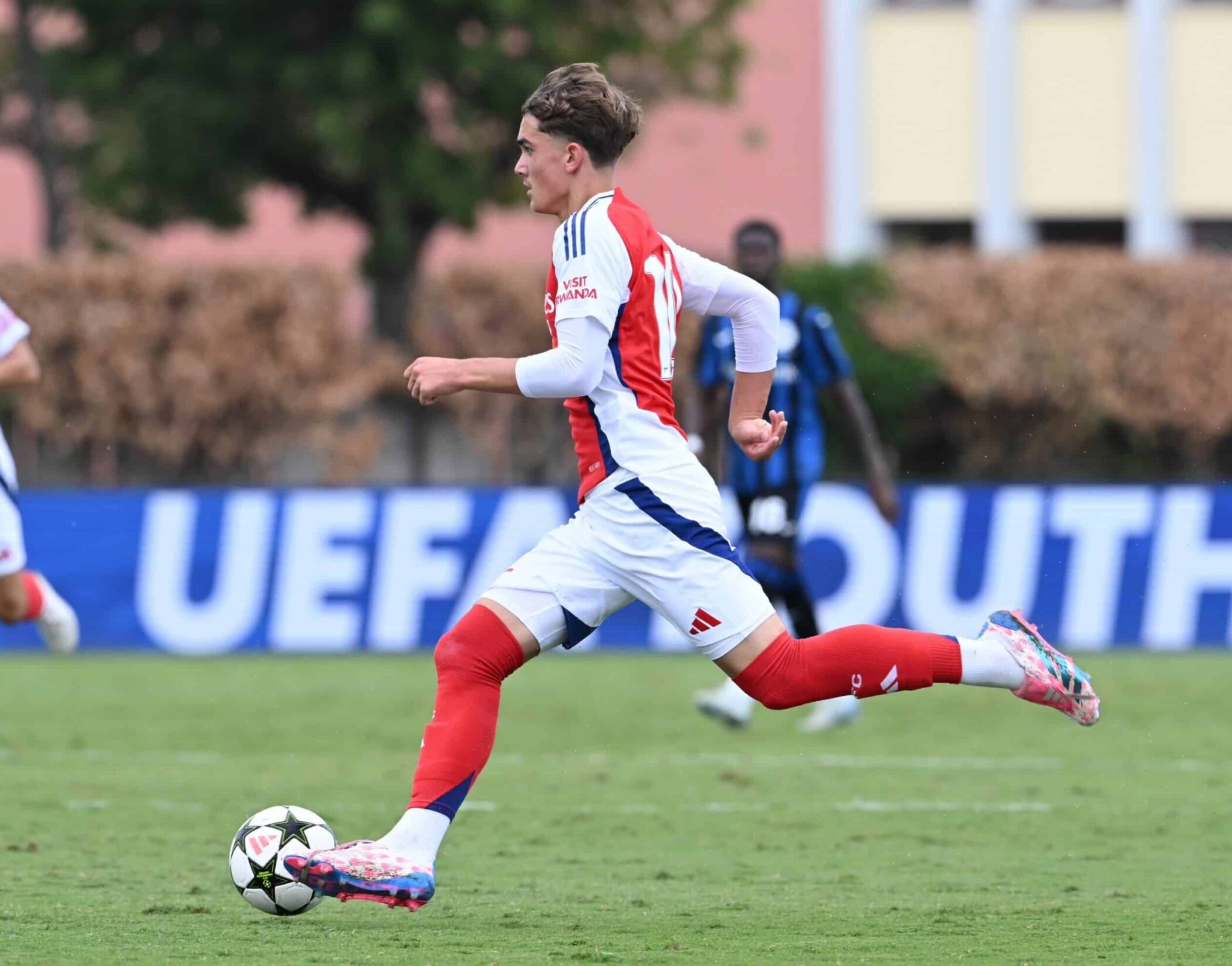 A footballer in a red and white kit runs with the ball on a grassy pitch during a UEFA youth match, where Max breaks football record, with blurred players and a banner visible in the background.