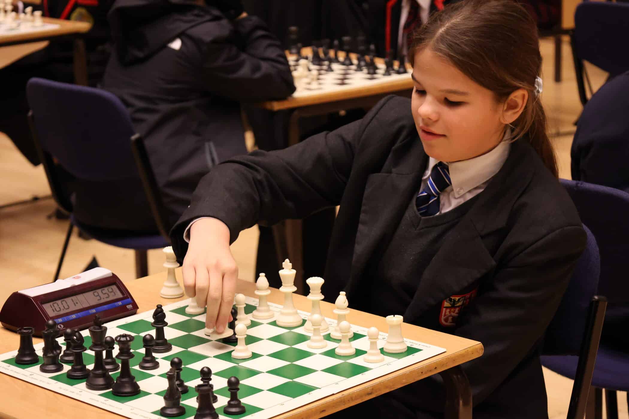 A young girl in a school uniform, sitting at a table, playing chess and moving a white chess piece on the board. Other chess games are taking place in the background.