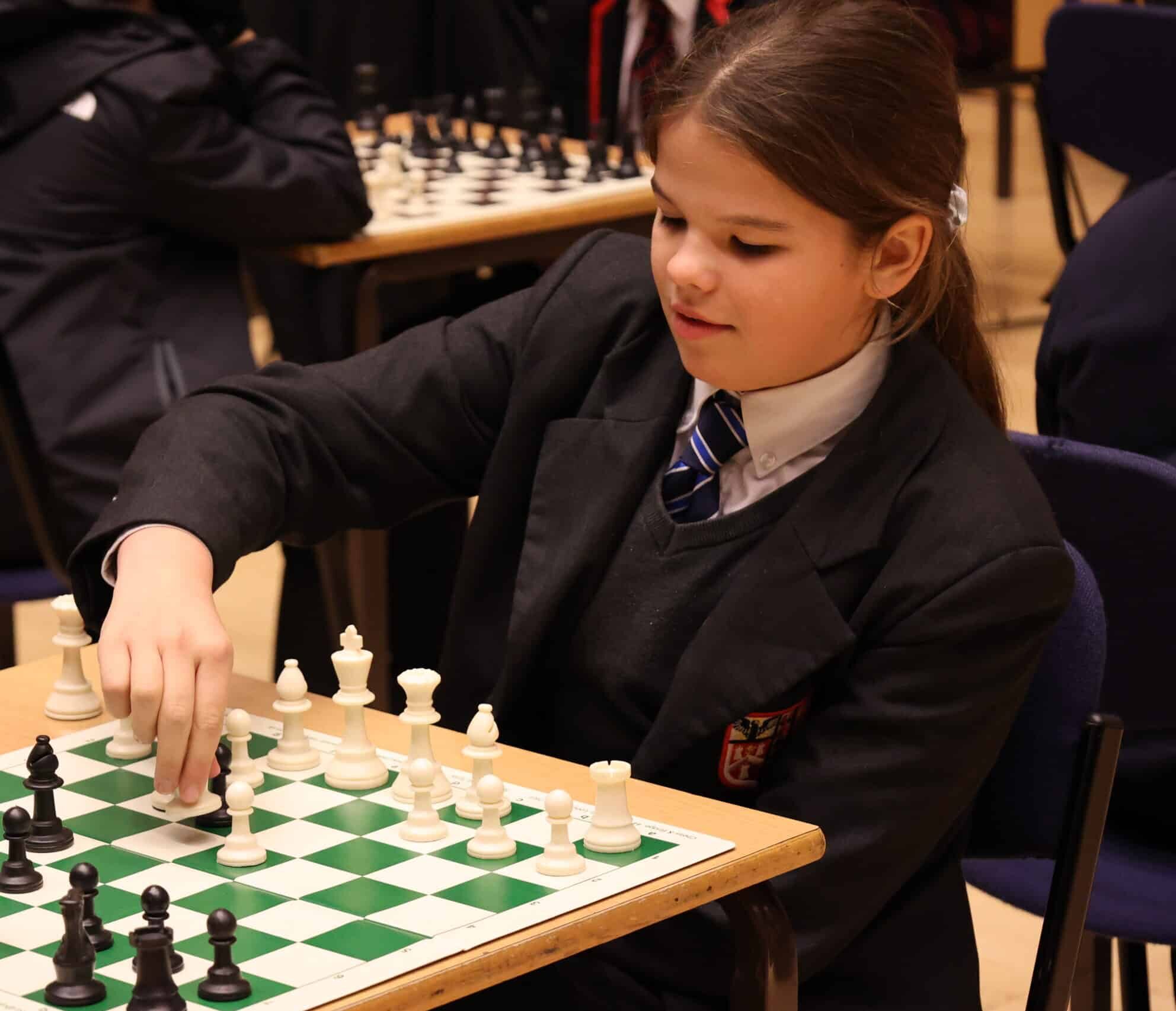A young girl in a school uniform, sitting at a table, playing chess and moving a white chess piece on the board. Other chess games are taking place in the background.
