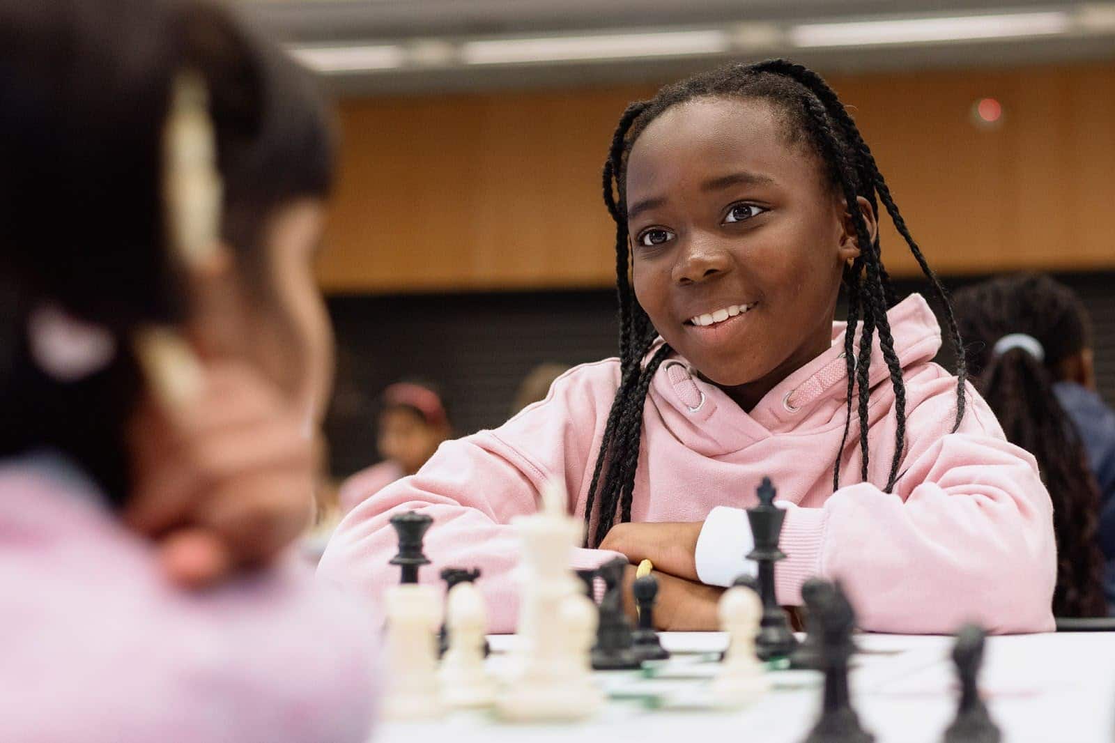 A young girl in a pink jumper smiles and sits at a table opposite another child, playing a game of chess indoors. Chess pieces and a blurred background are visible.