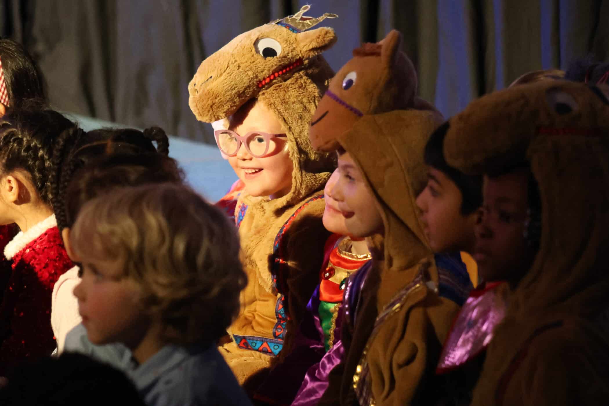 A group of children sit closely together, some wearing camel costumes and others in colourful outfits, smiling and looking towards the stage in a dimly lit room.