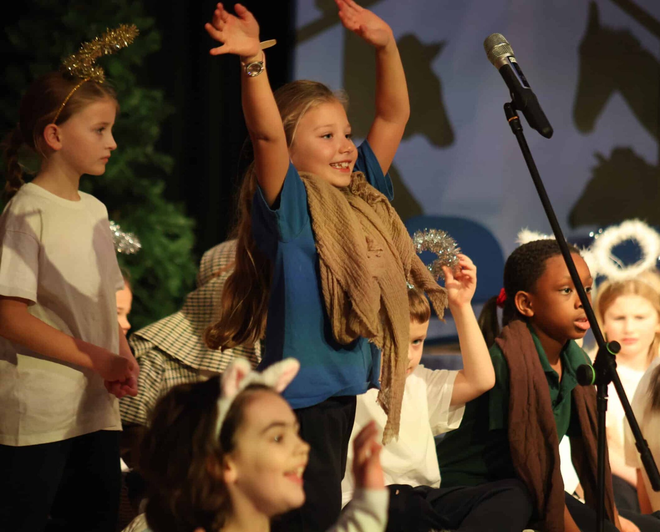 Children wearing costumes perform on stage. One girl stands with arms raised, smiling, while other children, some with halo and animal ear headbands, watch or participate. A microphone is visible in front of them.