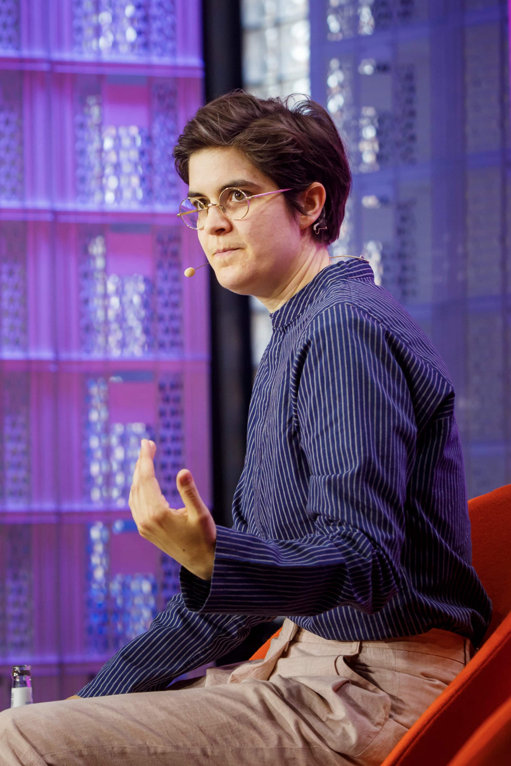 A person with short dark hair and glasses, wearing a striped shirt and beige trousers, sits in a red chair, gesturing while speaking on stage with a headset microphone about support and awareness for charities. The background features a purple geometric pattern.
