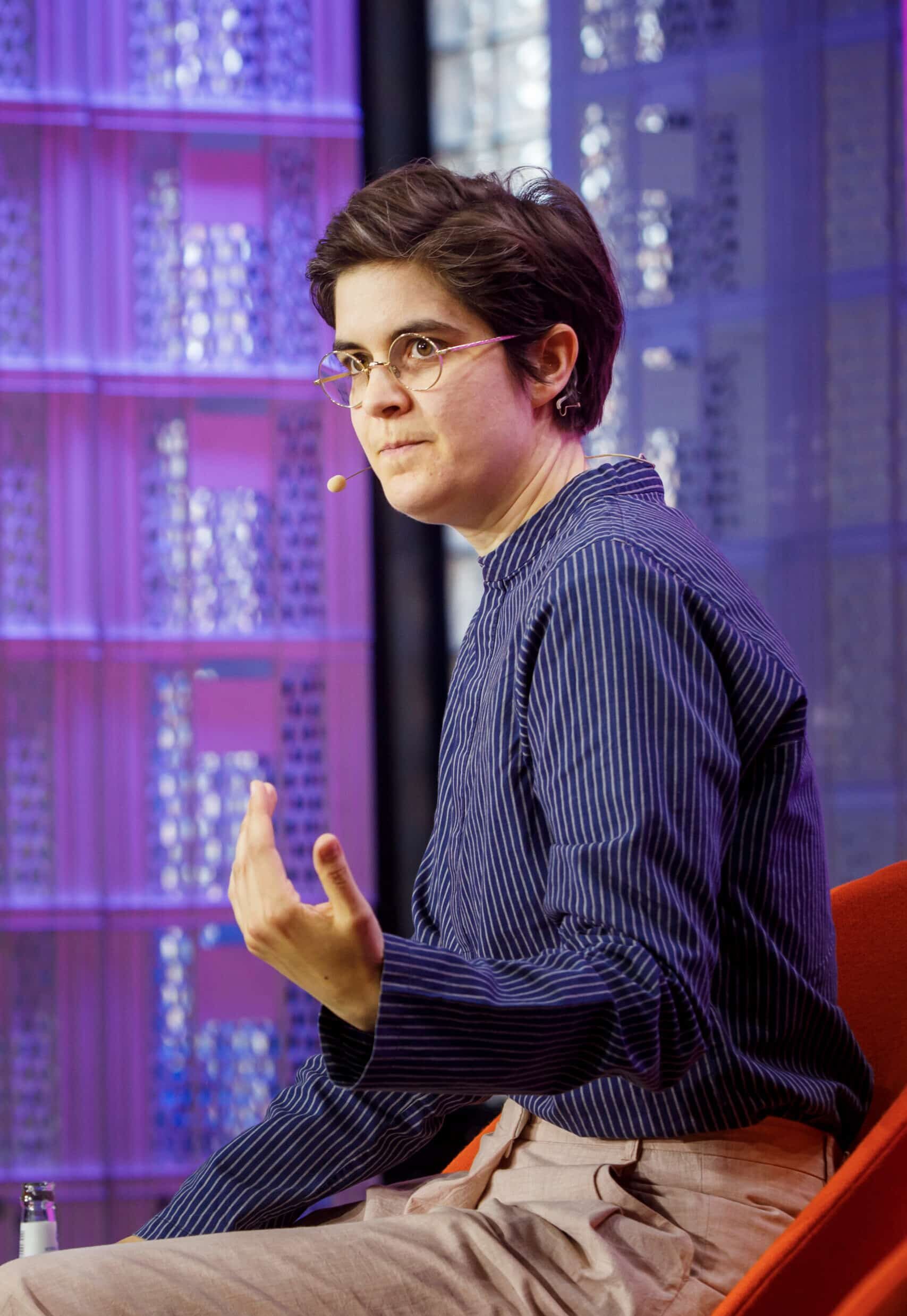 A person with short dark hair and glasses, wearing a striped shirt and beige trousers, sits in a red chair, gesturing while speaking on stage with a headset microphone about support and awareness for charities. The background features a purple geometric pattern.