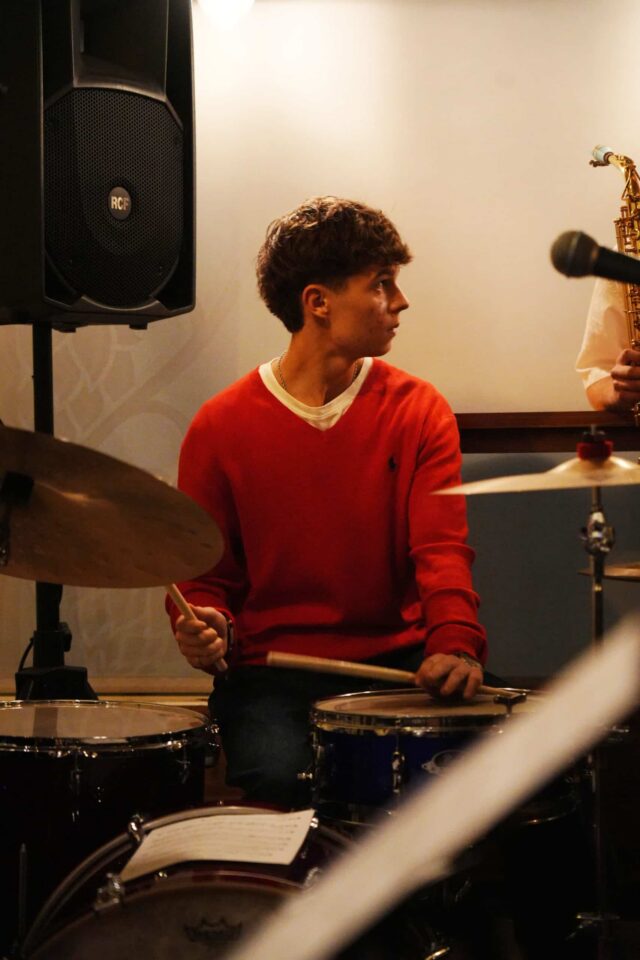 A young man in a red jumper plays the drums, looking to his right. A saxophone and a speaker are visible in the background, suggesting a live music performance.