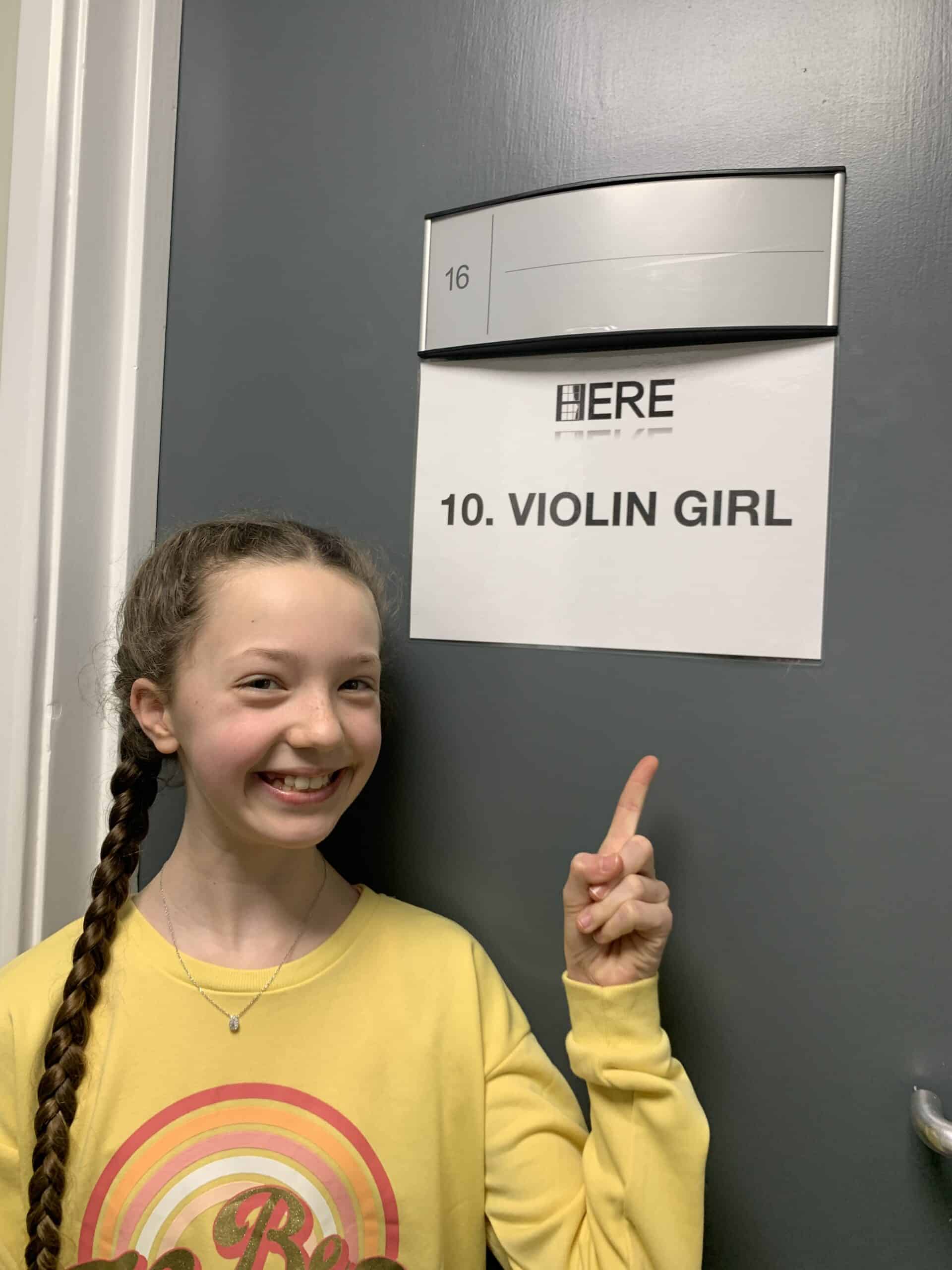 A smiling girl with a long plait, wearing a yellow shirt, points at a door sign that reads 10. VIOLIN GIRL beneath a larger, partially covered sign.