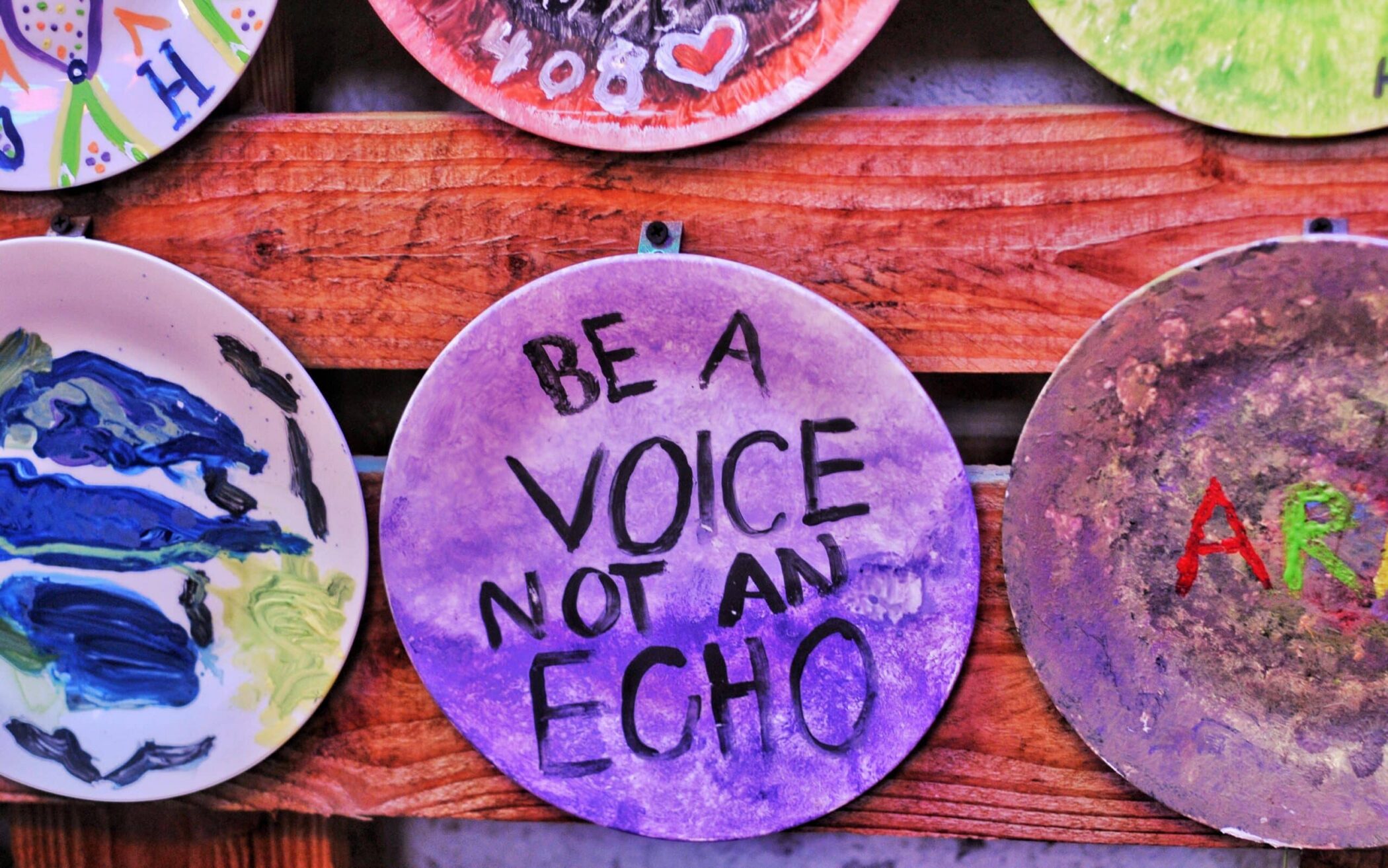 A round ceramic plate painted purple with the words Be a VOICE not an ECHO displayed on a wooden wall among other decorated plates.