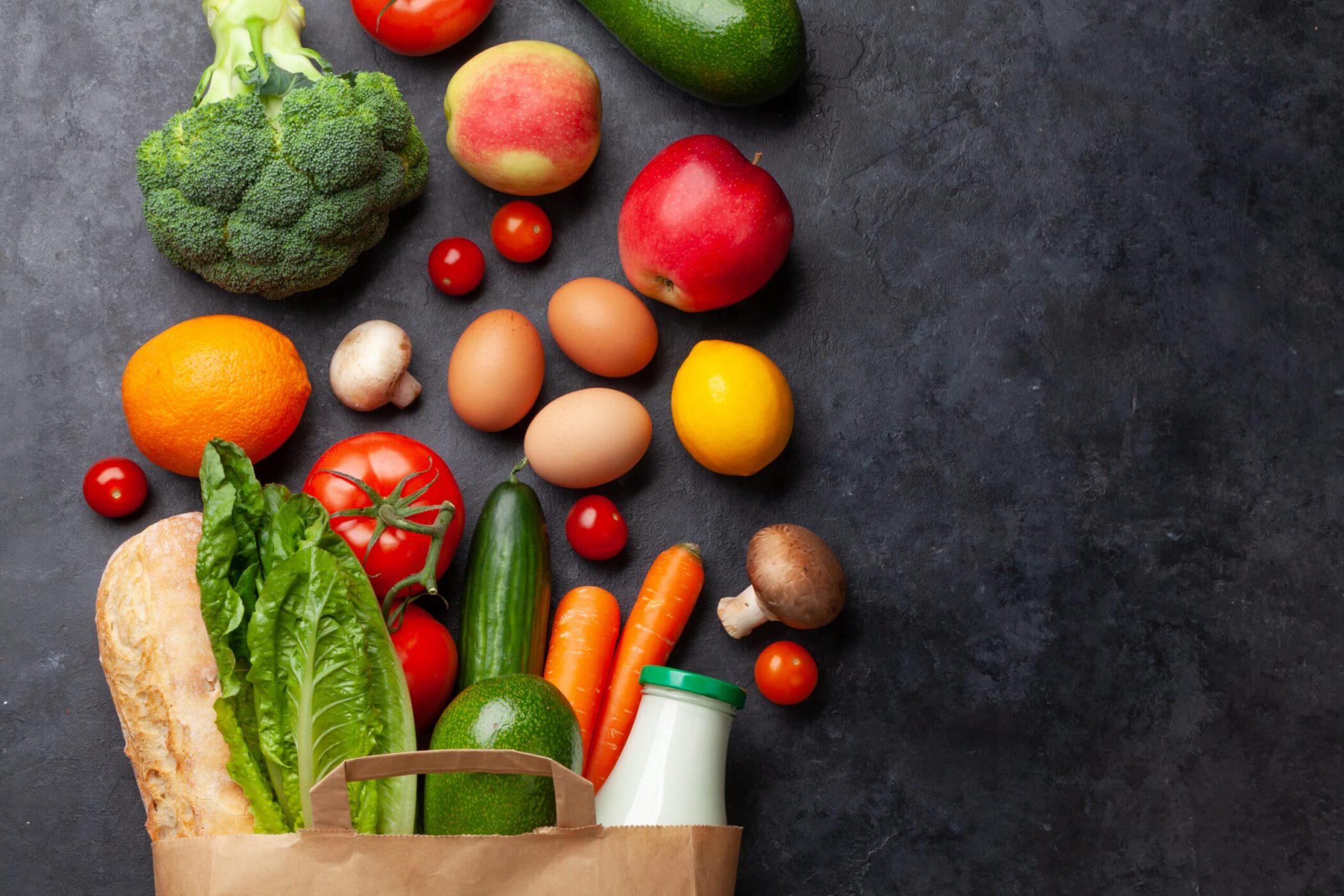 A variety of fresh groceries, including broccoli, apples, oranges, tomatoes, carrots, lettuce, eggs, mushrooms, cucumber, a baguette, and a bottle of milk, are arranged on a dark surface with a paper bag.
