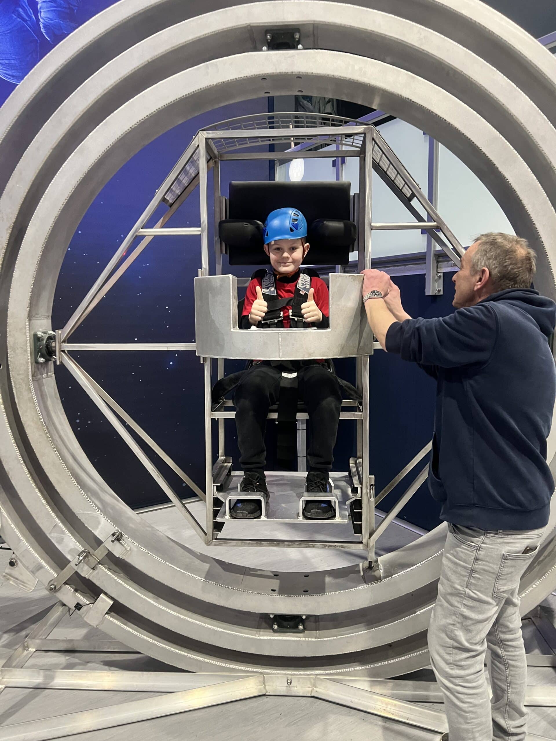 A child wearing a blue helmet sits in a metal gyroscope ride giving two thumbs up, while an adult in a hoodie stands next to the structure, preparing the ride.
