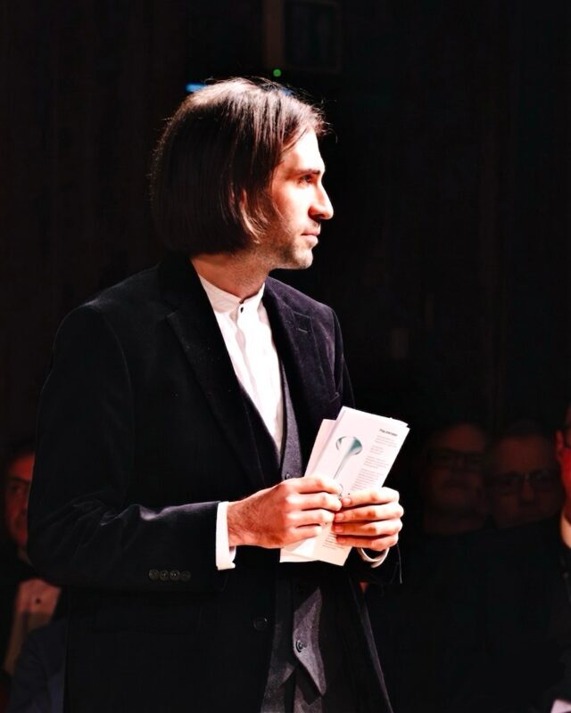 A man with dark hair and a beard, wearing a white shirt, smiles whilst holding a violin and bow. The background is blurred, with indistinct figures and lights. The black-and-white photo captures Grig Cuciuc’s warm presence.