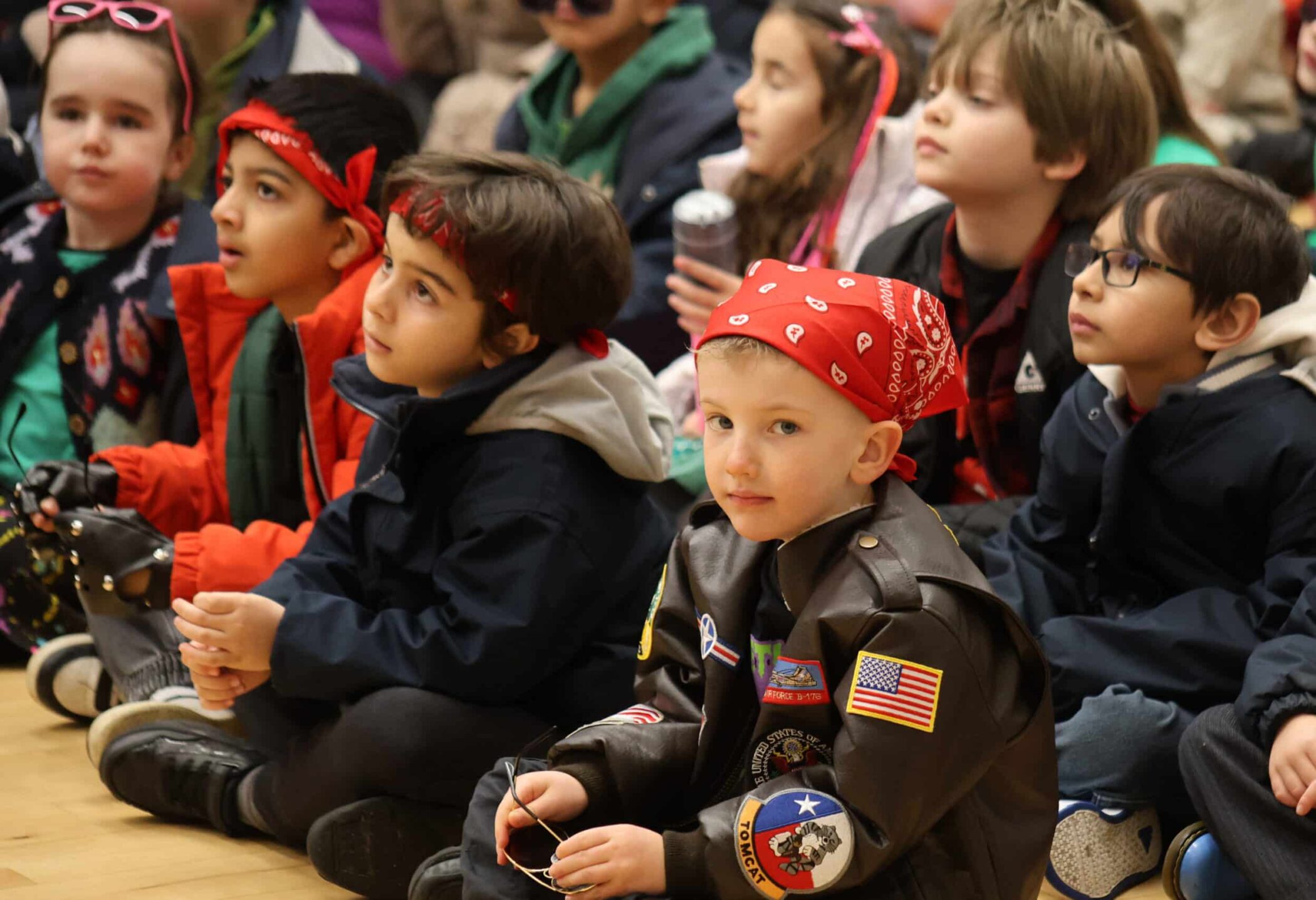 A group of young children sit on the floor, dressed in colourful outfits and bandanas, attentively watching something off-camera. One child in front wears a leather jacket with patches and holds sunglasses.