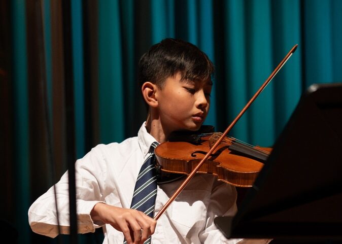 A young boy in a white shirt and striped tie plays the violin on stage at the Brentwood International Music Competition, focused on his performance, with a blue curtain in the background.
