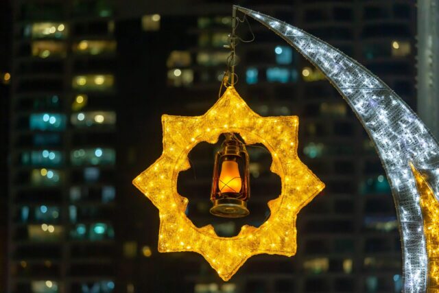 A glowing yellow star-shaped light frame with a lantern hanging inside, next to a lit crescent moon decoration, set against a background of tall buildings at night.