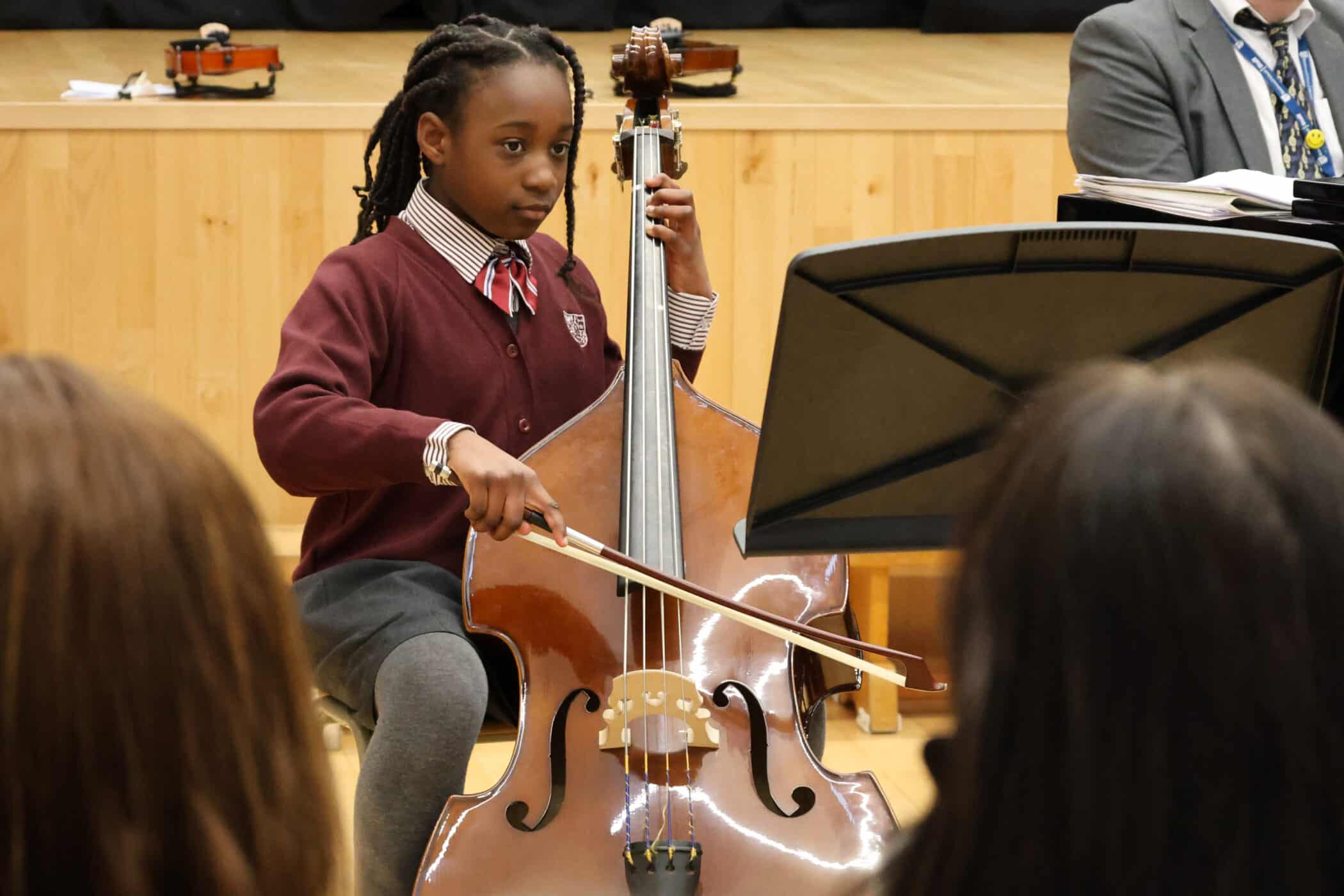A young student in a school uniform plays a double bass with a bow, focusing on sheet music during a performance. Several people watch in the audience, and small violins are visible in the background.