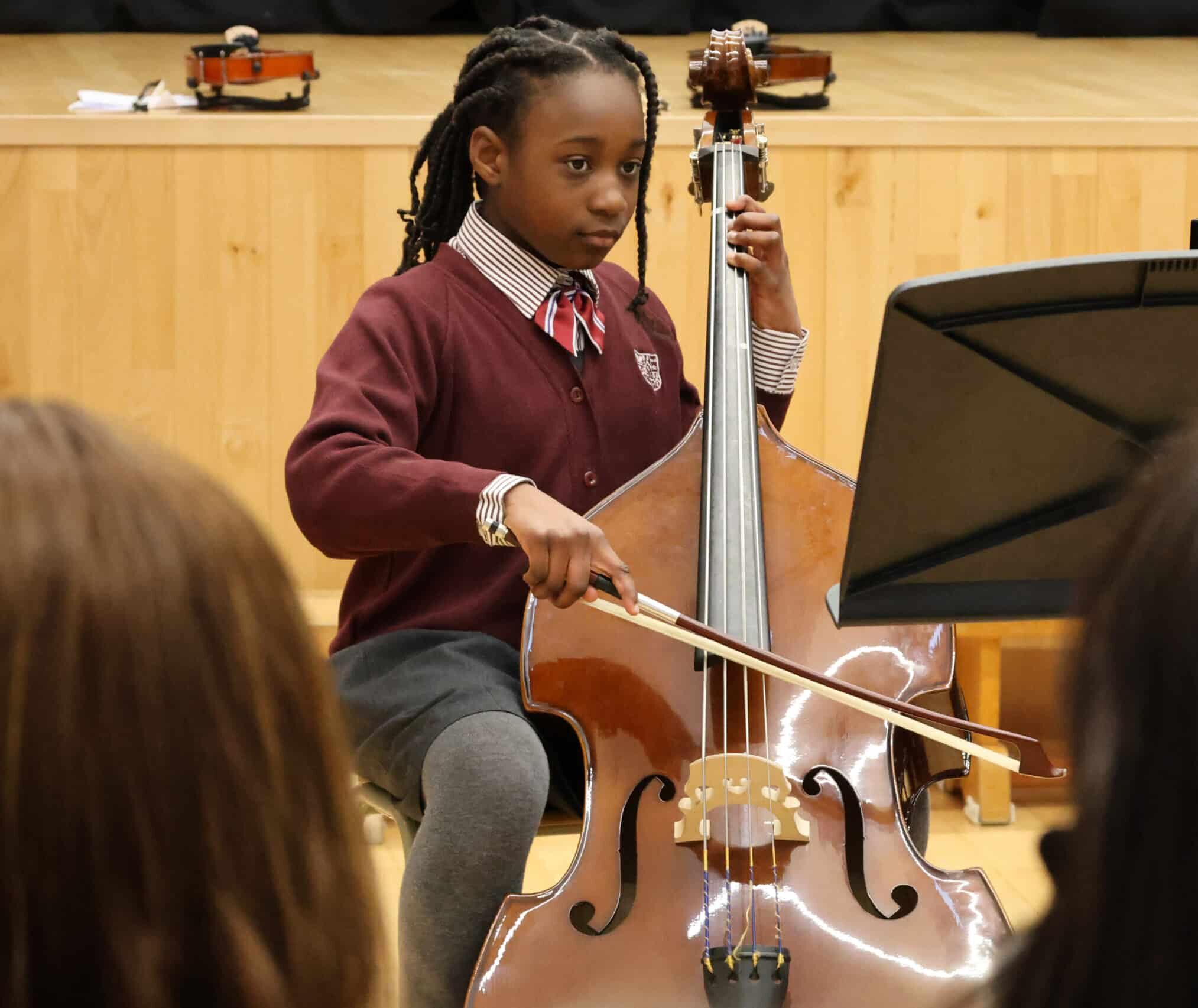A young student in a school uniform plays a double bass with a bow, focusing on sheet music during a performance. Several people watch in the audience, and small violins are visible in the background.