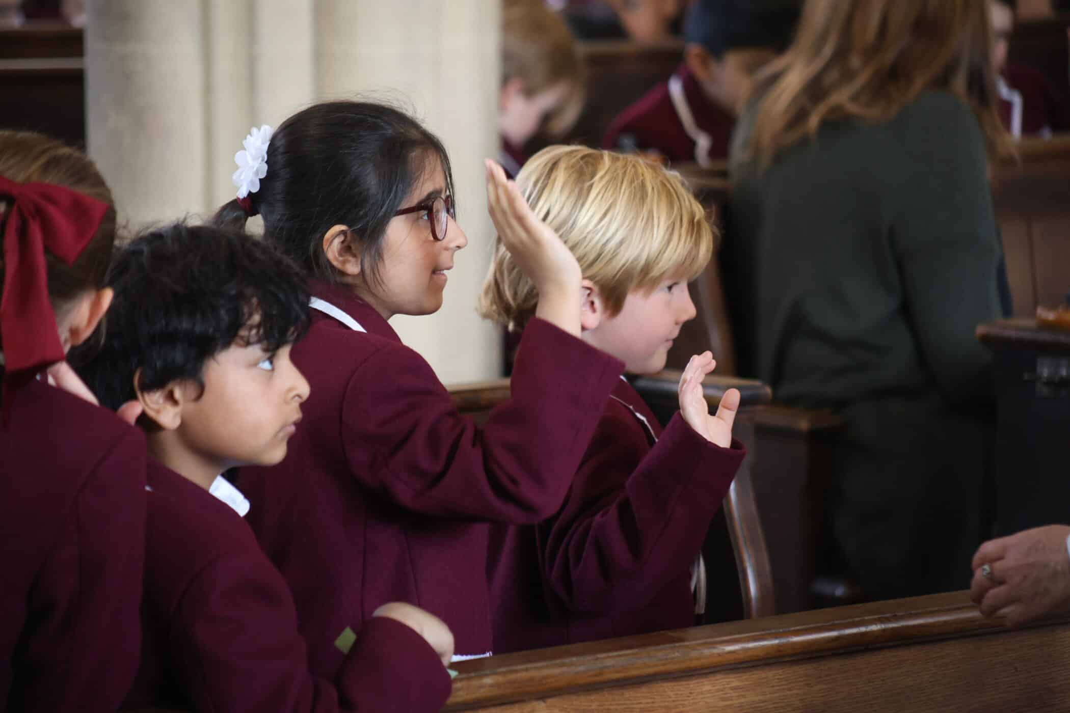 Three young children in school uniforms sit in a row inside a church or assembly hall. One girl with glasses raises her hand, whilst the other two children sit attentively. All appear focused on something out of frame.