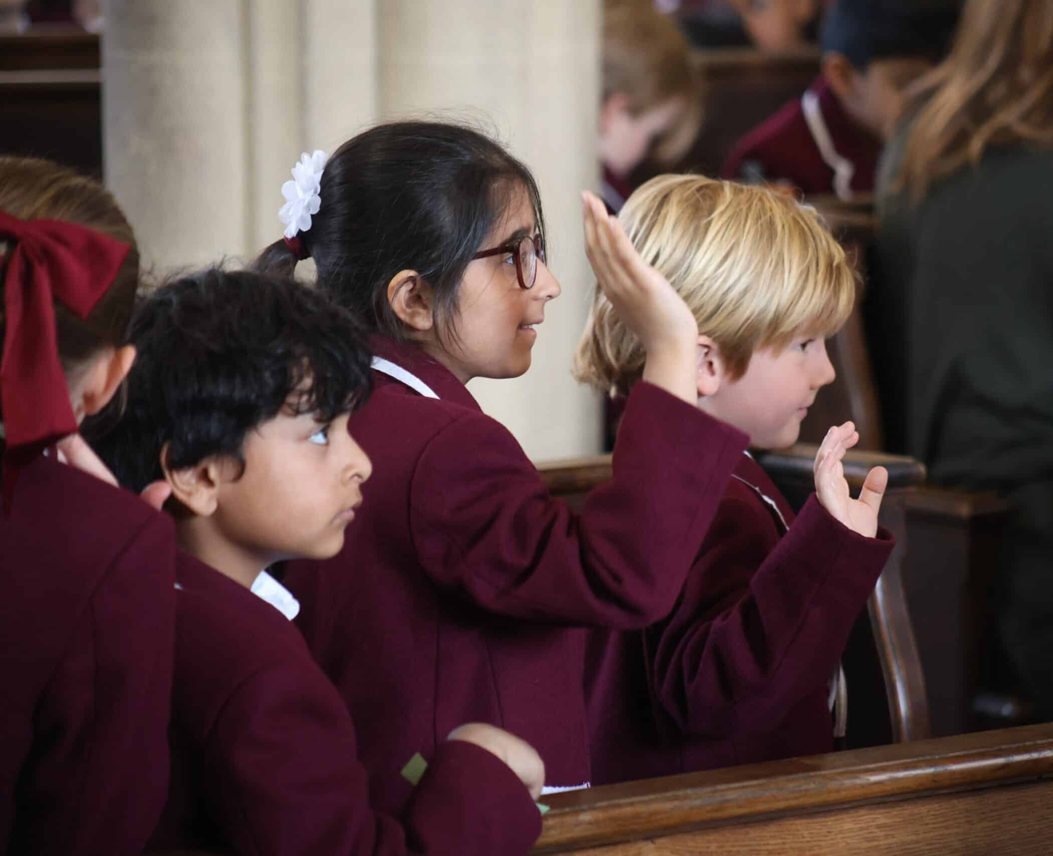 Three young children in school uniforms sit in a row inside a church or assembly hall. One girl with glasses raises her hand, whilst the other two children sit attentively. All appear focused on something out of frame.