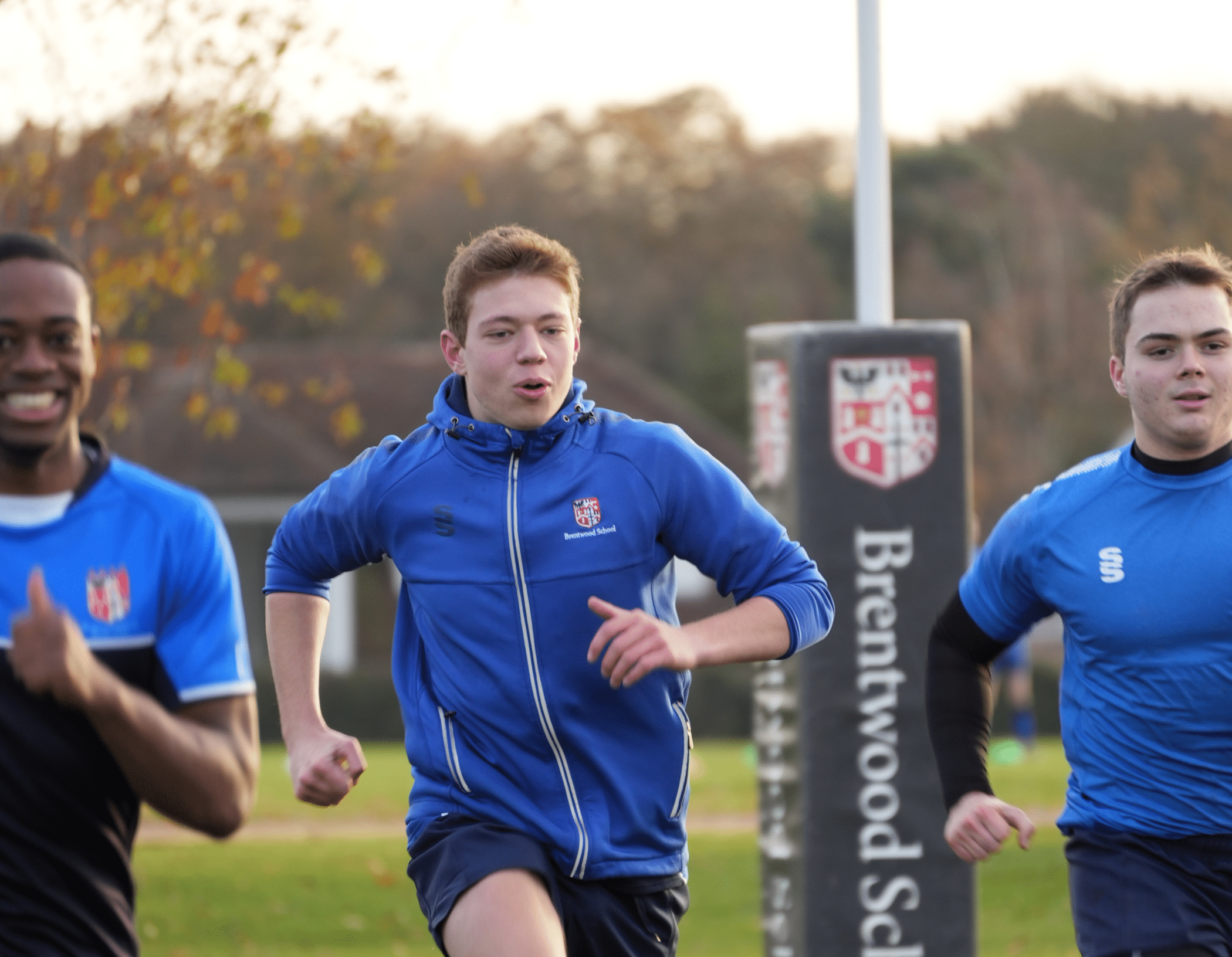 Three young men in blue sports kit are running outdoors on a grassy pitch at this leading IB Boarding School. A rugby post with the Brentwood School logo is visible in the background, with trees in the distance.