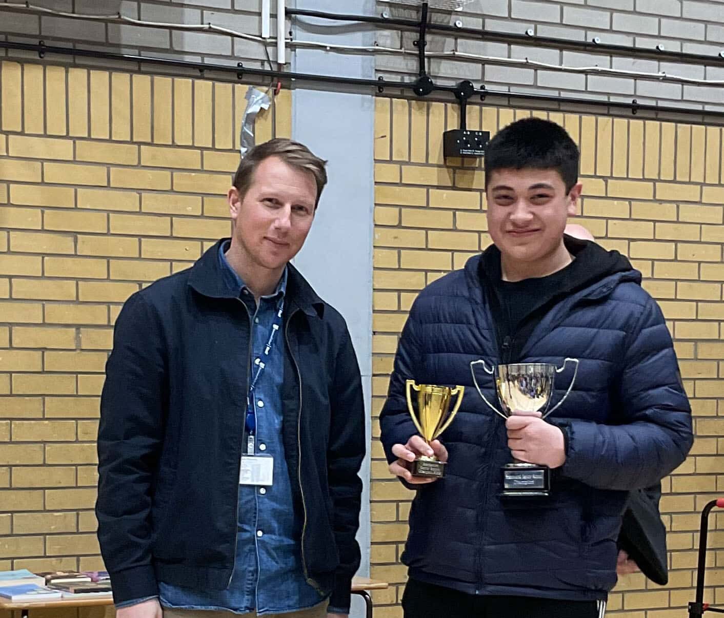 Two men stand indoors against a yellow brick wall. The man on the right is smiling and holding two gold trophies, whilst the man on the left stands beside him, wearing a lanyard and dark jacket.