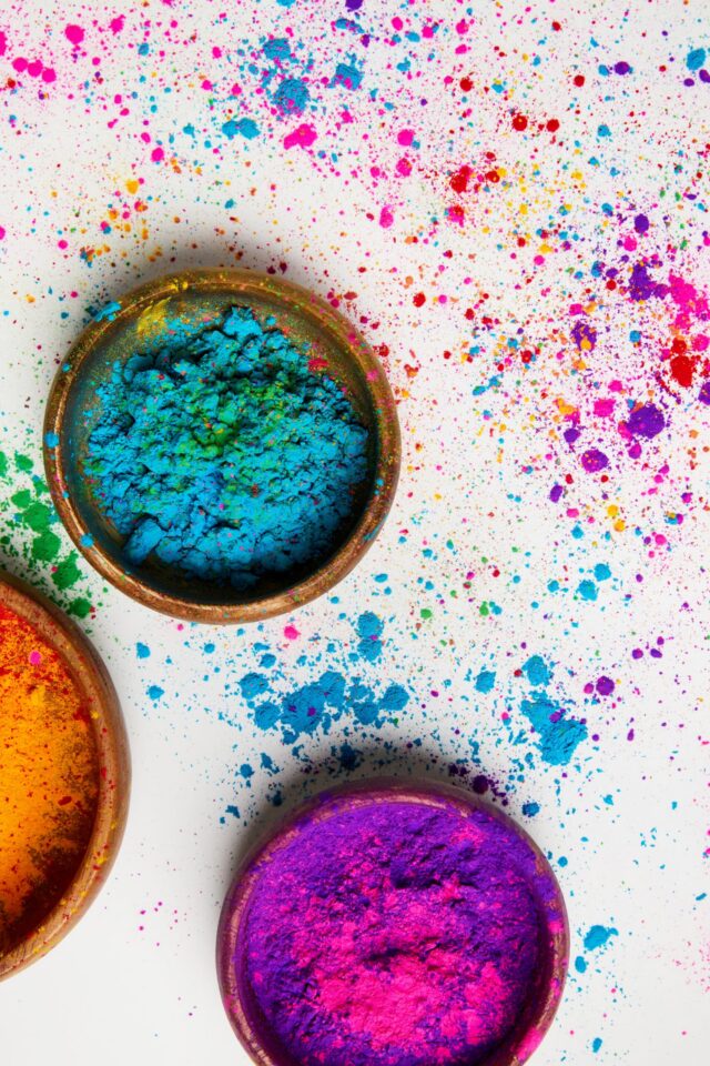 Bowls filled with colourful powder—blue, purple, and orange—sit on a white surface, with vibrant powder splattered around them, suggesting a festive or artistic celebration like Holi.