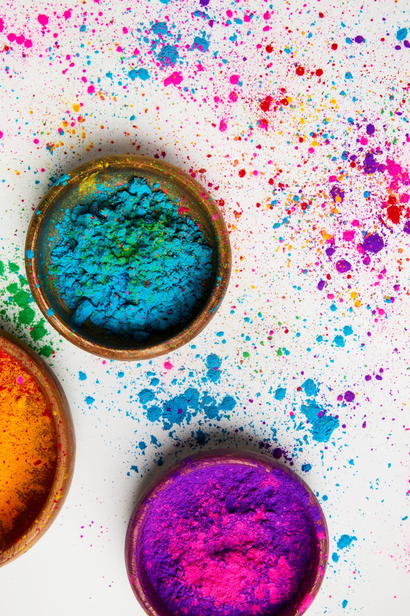 Bowls filled with colourful powder—blue, purple, and orange—sit on a white surface, with vibrant powder splattered around them, suggesting a festive or artistic celebration like Holi.