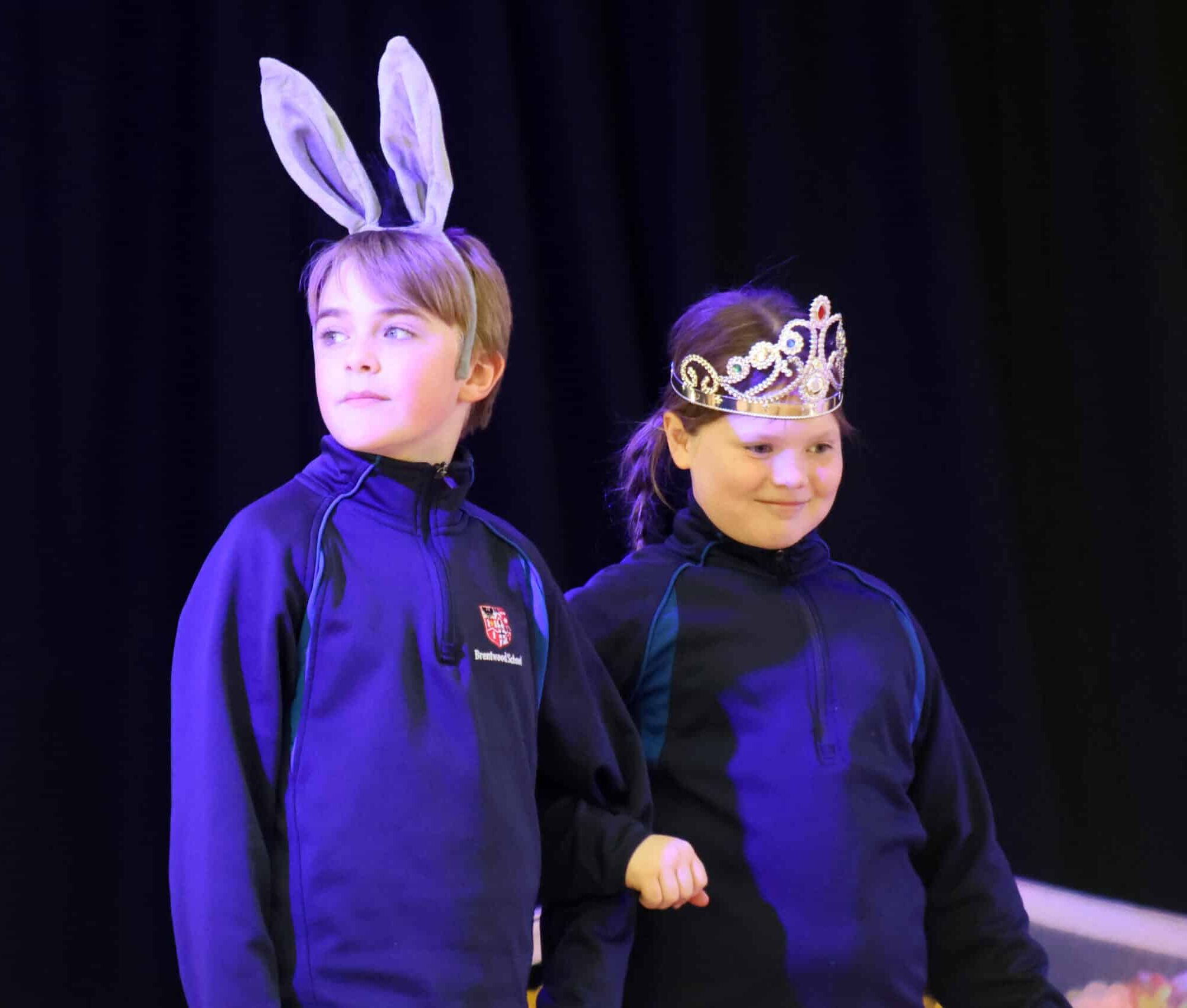 Two children in dark outfits stand on stage; one wears grey bunny ears, and the other wears a sparkly tiara. Both look towards the audience with calm expressions against a dark background.