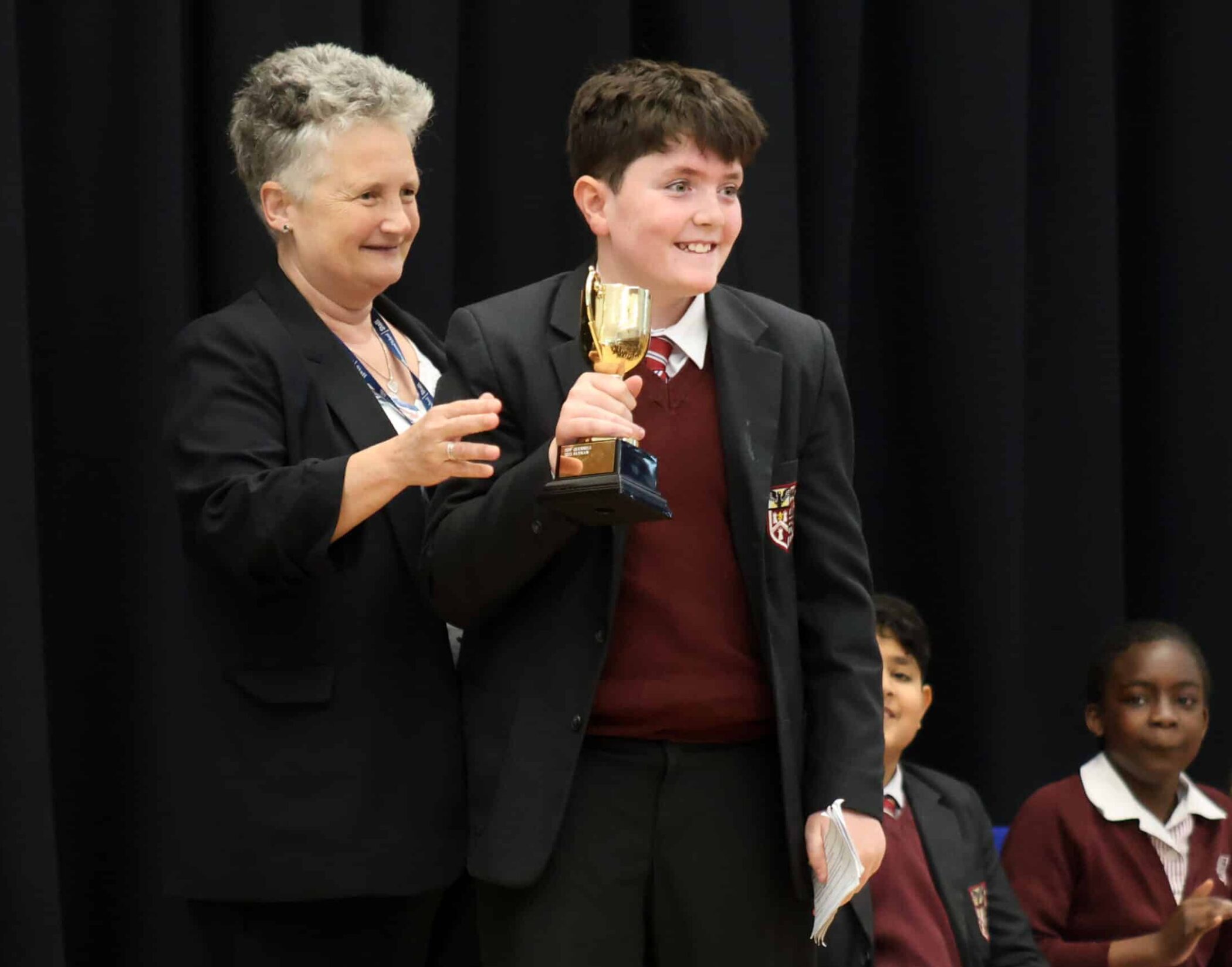 A smiling boy in a school uniform holds a trophy whilst standing next to a woman. Two other children in uniforms are seated in the background against a black curtain.