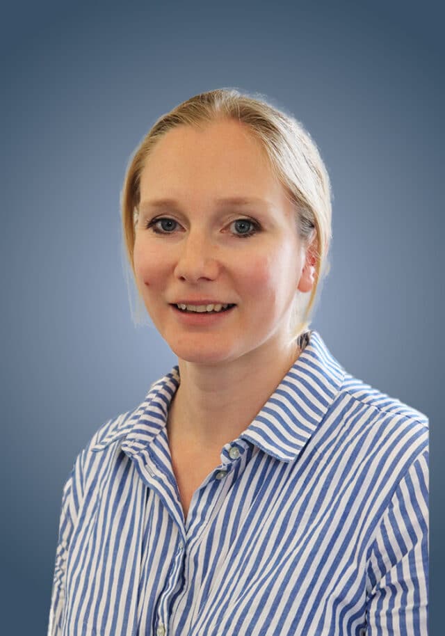 A woman with blonde hair, wearing a blue and white striped shirt, smiles at the camera against a plain blue gradient background, embodying the approachable professionalism of a Director of IB.