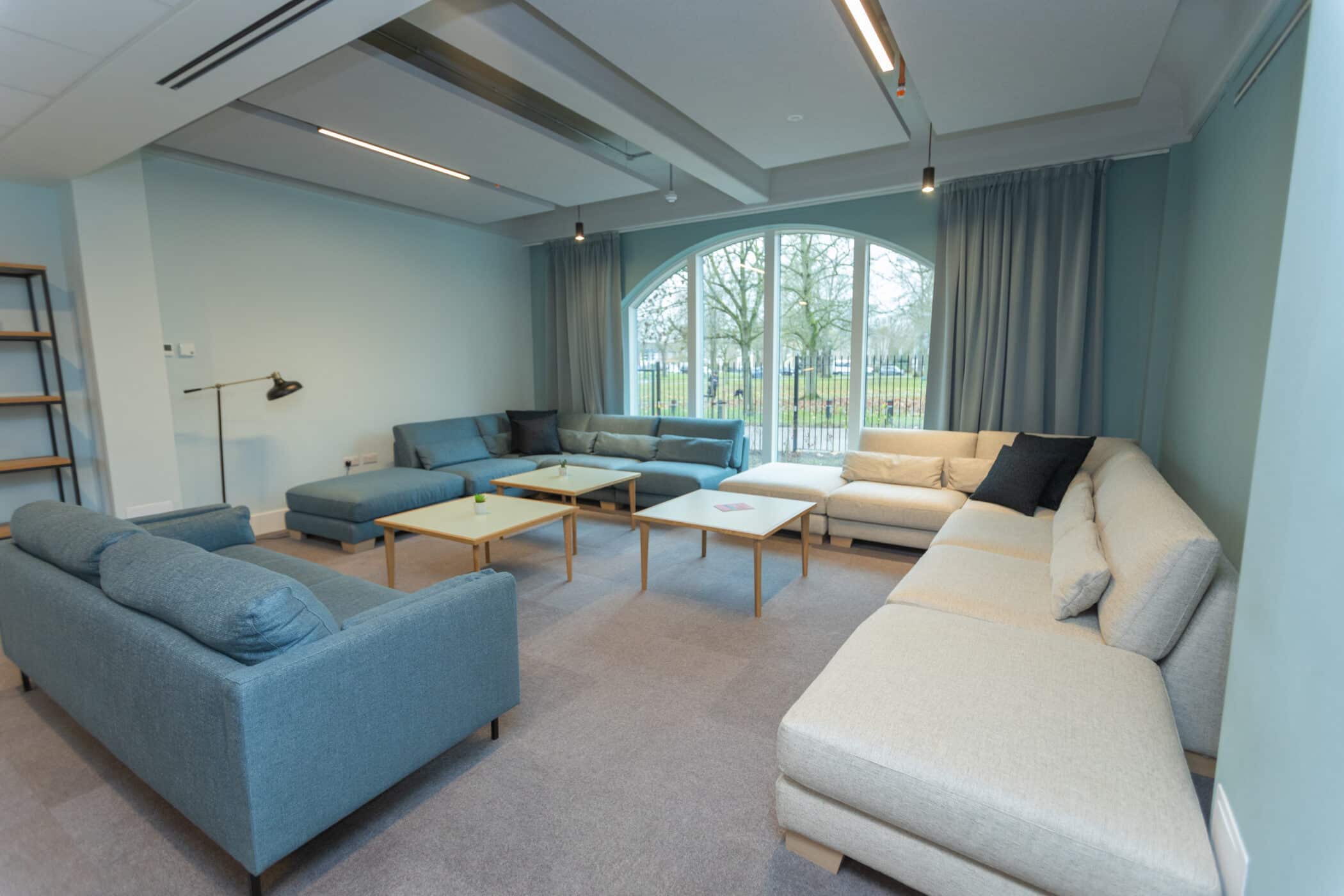A cosy living room at the New Boarding House features pastel green walls, two beige sofas, a blue sofa, light wooden tables, board games, a window with curtains, and a large TV displaying this morning.