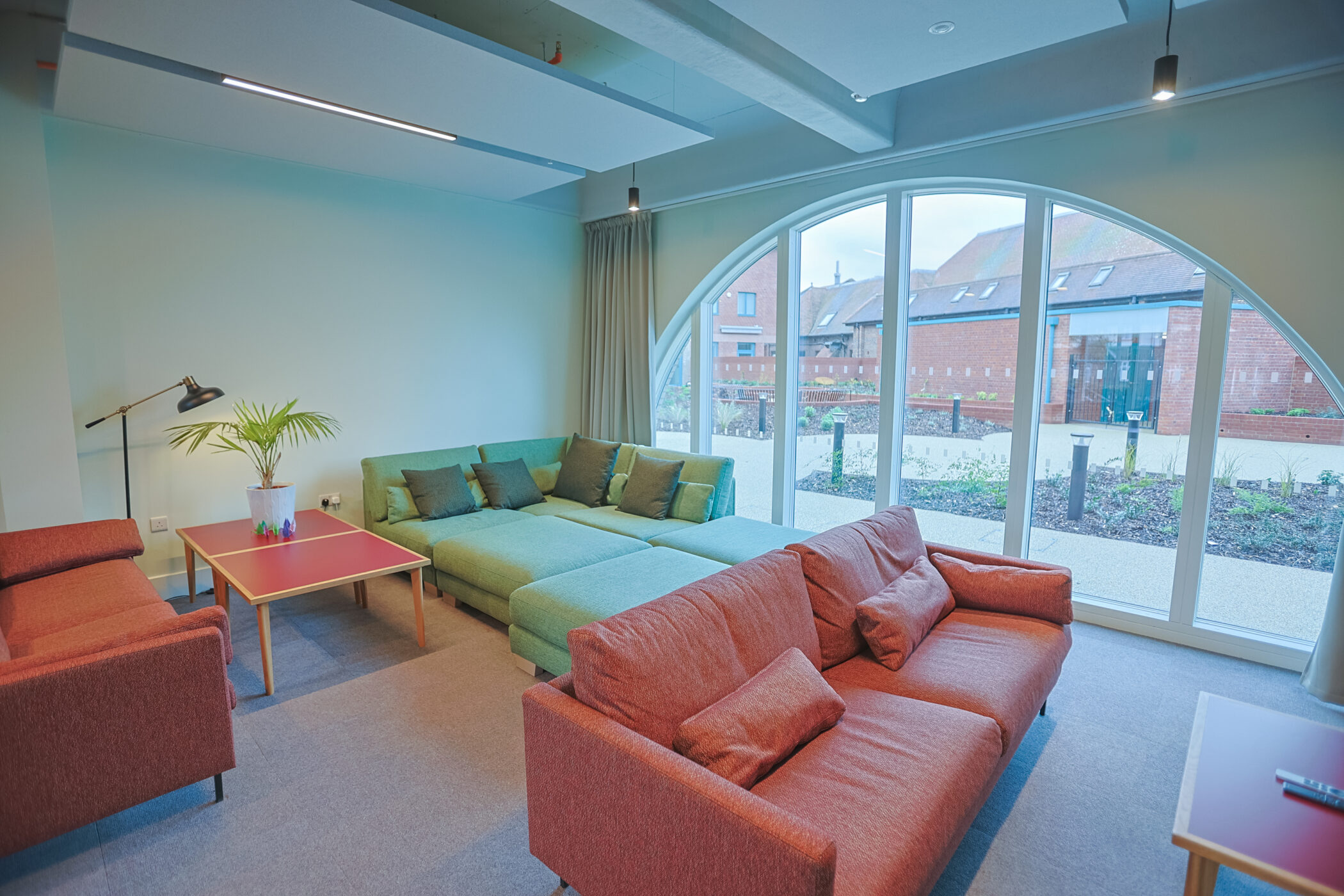 Modern lounge area in the New Boarding House features a large arched window, multiple sofas in green and brown tones, several coffee tables, and a wall-mounted television; enjoy views of brick buildings outside the window.