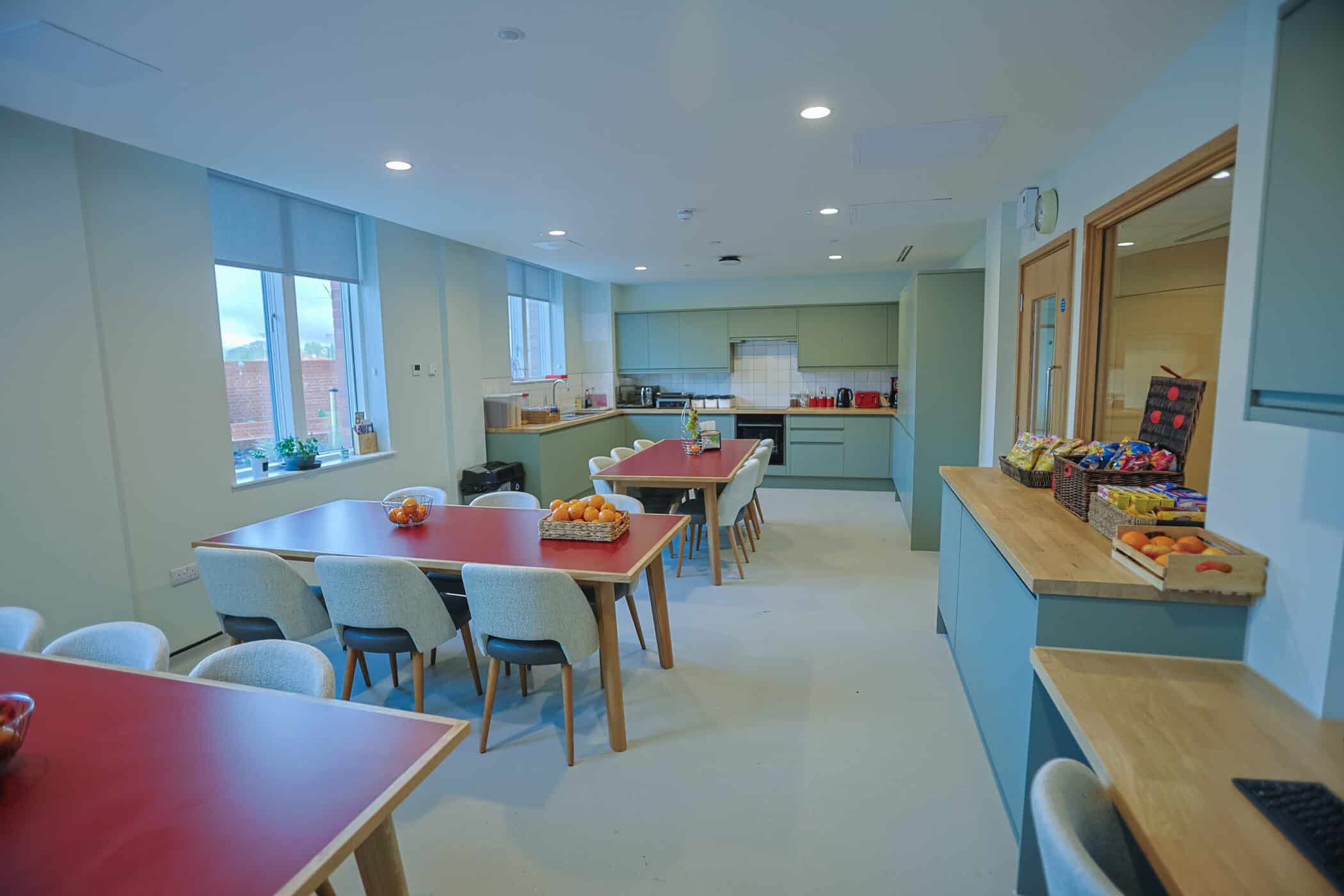 A modern communal kitchen in the New Boarding House features light grey cupboards, red tabletops, beige upholstered chairs, and large windows letting in natural light. The space is clean and neatly organised.