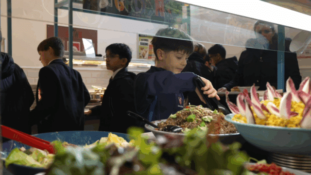 A young boy serves himself salad from a buffet, with other pupils queued up behind him. Brightly coloured vegetables are displayed in large bowls in the foreground.