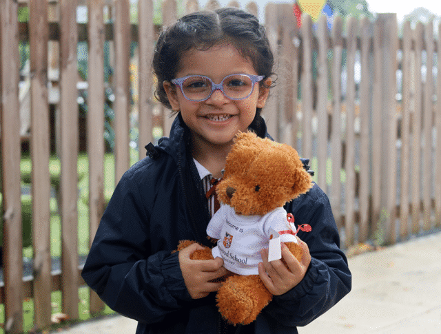 A young girl with glasses smiles whilst holding a teddy bear dressed in a white shirt. She is wearing a dark jacket and standing outside near a wooden fence, adding to the pastoral charm of the scene.