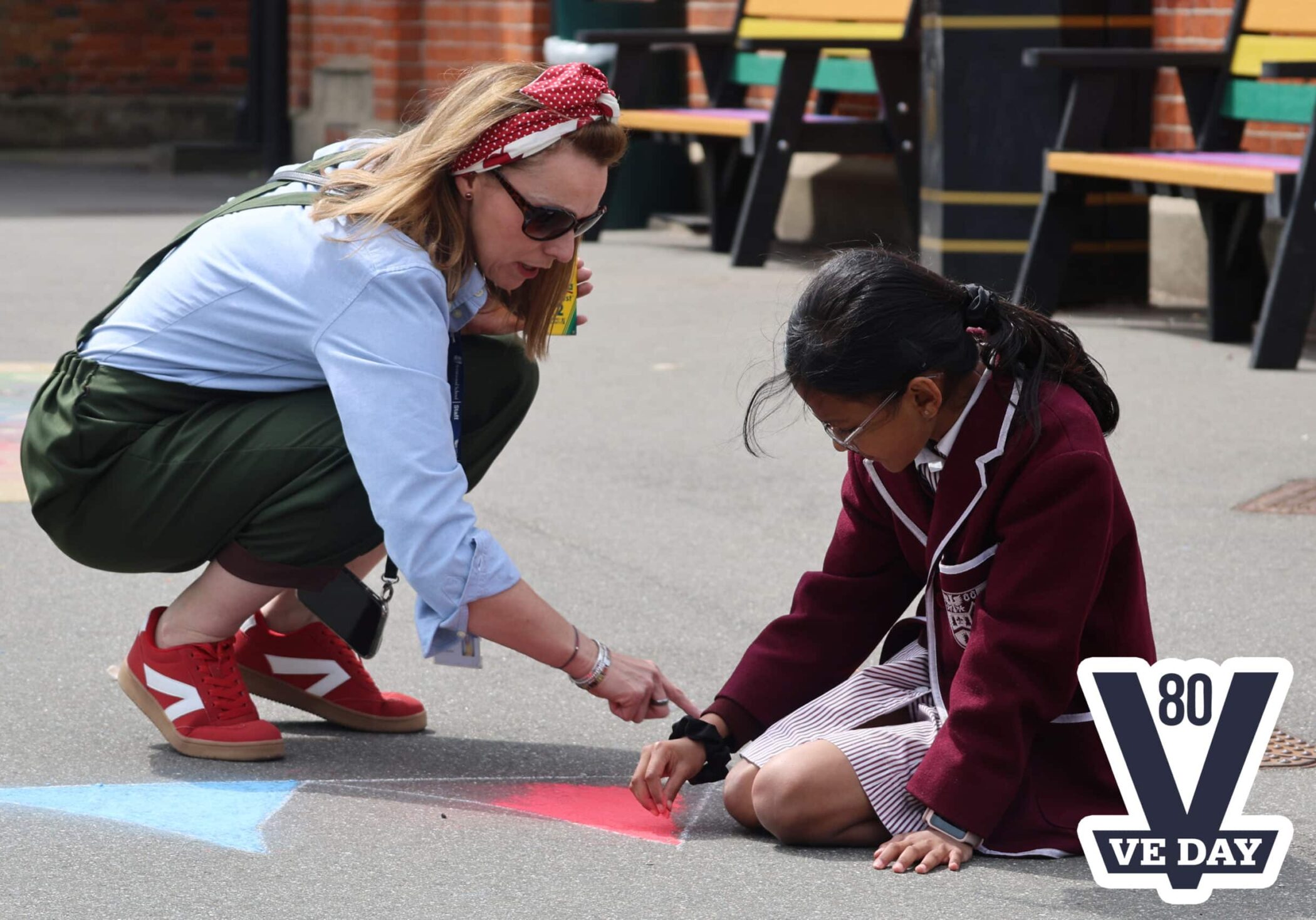 A woman kneels on a playground pointing at chalk drawings whilst a girl in a school uniform draws on the ground. A VE DAY 80 logo is in the corner.