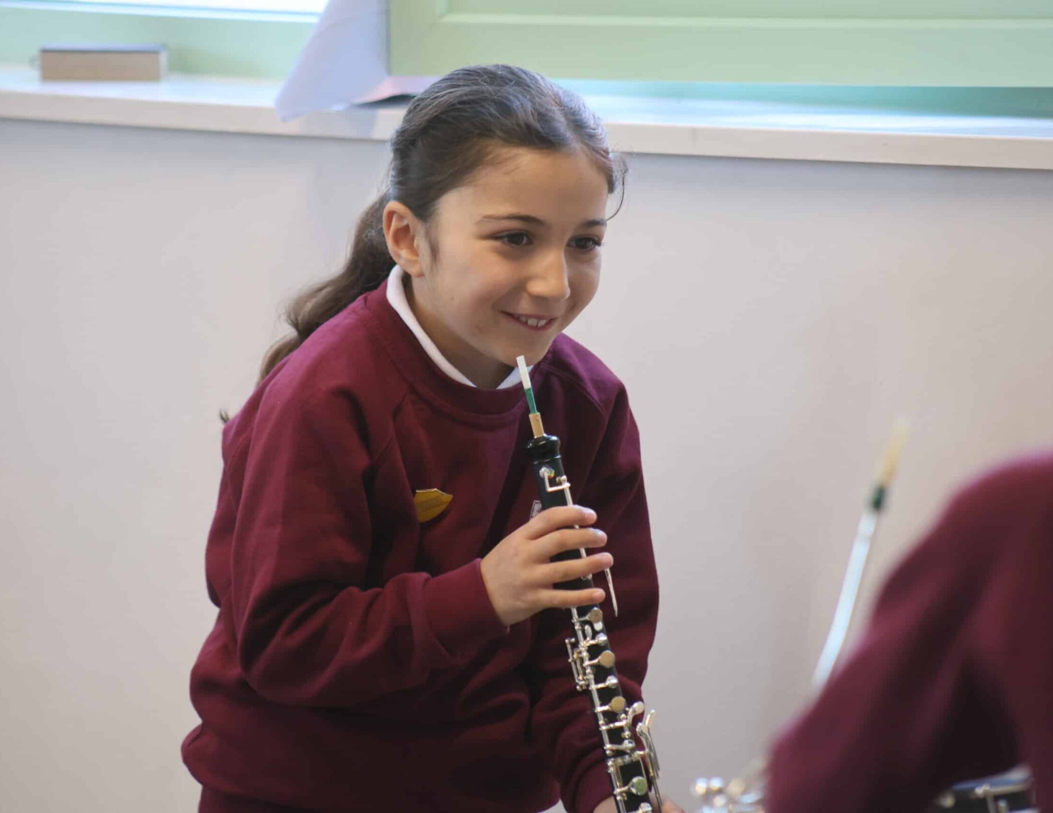 A young girl in a maroon school uniform holds and plays an oboe, smiling as she looks at another person out of shot in a classroom setting.