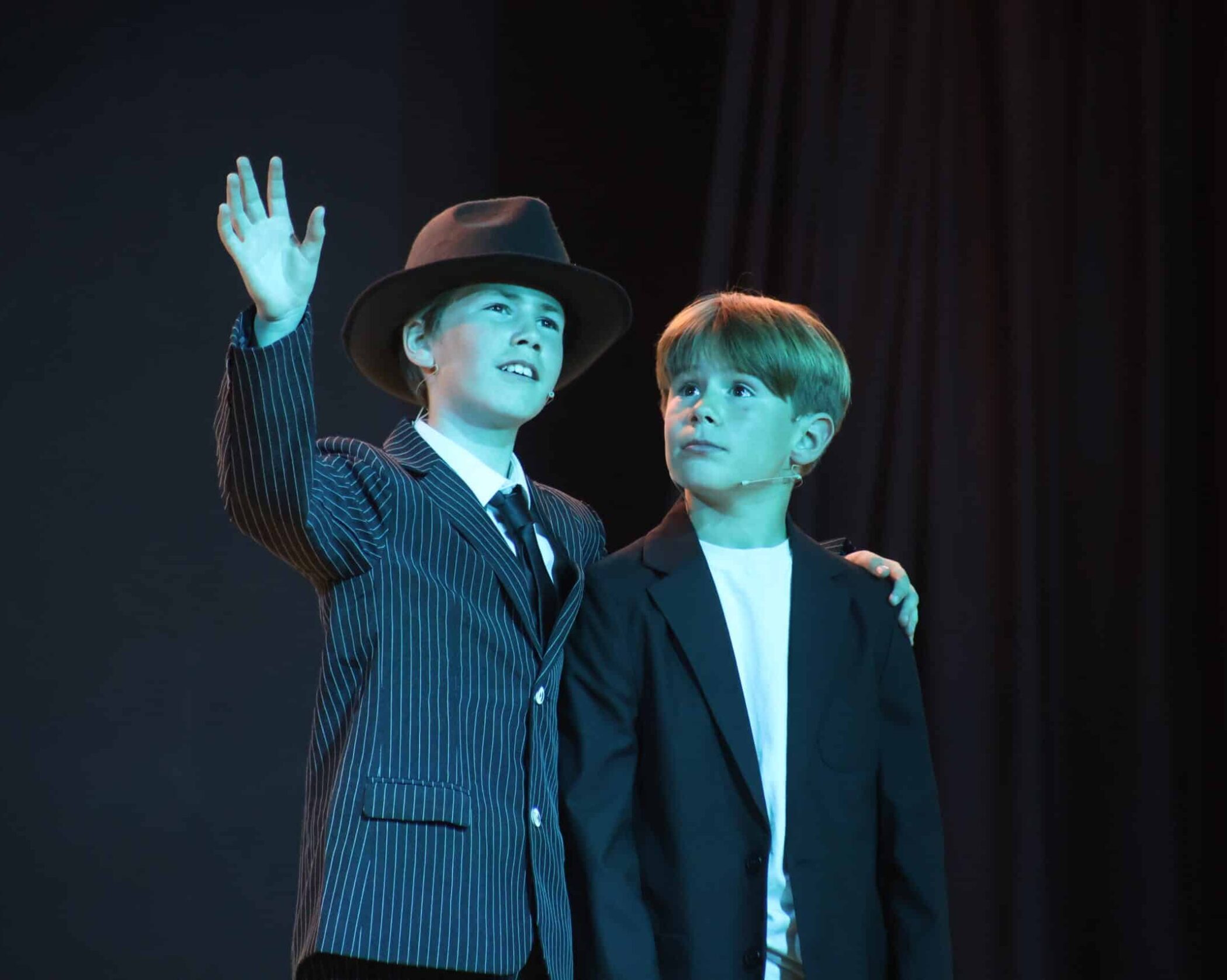 Two boys stand on stage in suits. One, wearing a pinstripe suit and hat, smiles and gestures with his hand raised. The other boy, in a plain dark jacket, looks at him with an attentive expression.