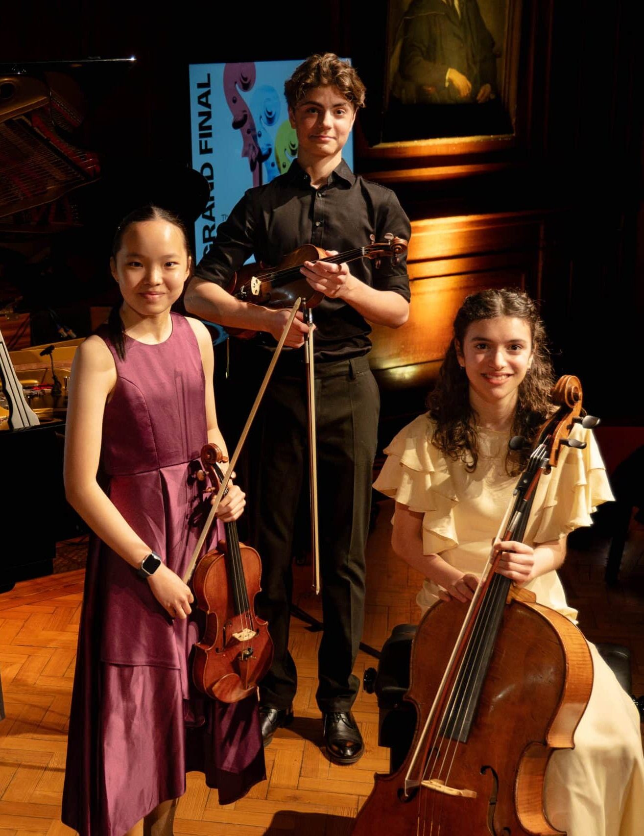 Three young musicians pose on a wooden stage at the Brentwood International Music Competition: a girl in a purple dress holding a violin, a boy in black with his violin and bow, and a girl in cream seated with her cello. A competition sign is visible behind them.