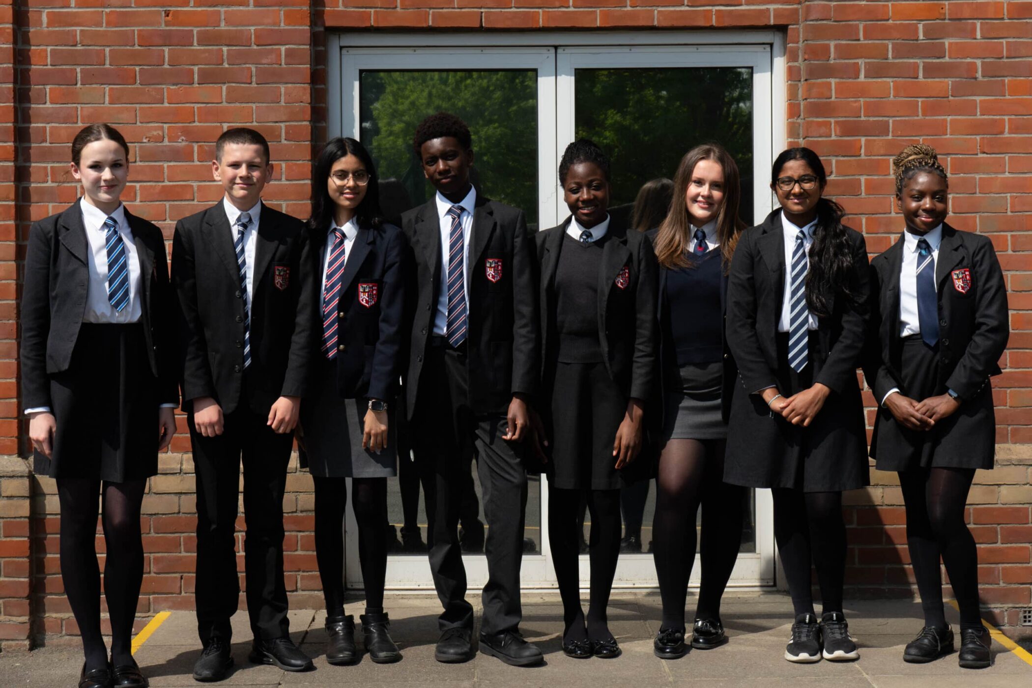 Eight students in school uniforms stand in a row outside a brick building with a glass door, smiling at the camera. The uniforms are black blazers, white shirts, and striped ties.