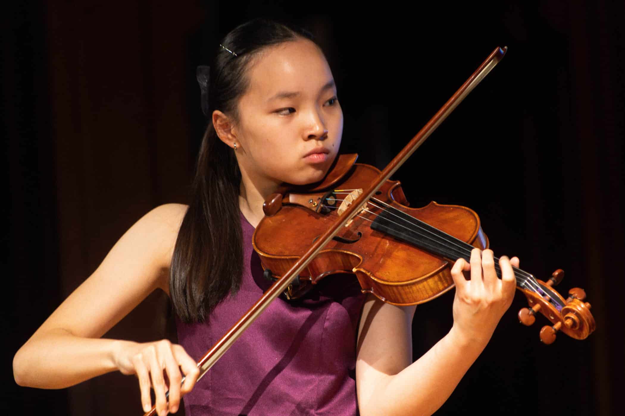 A young woman with long dark hair plays a violin on stage at the Brentwood International Music Competition, wearing a sleeveless purple top and looking focused as she performs.