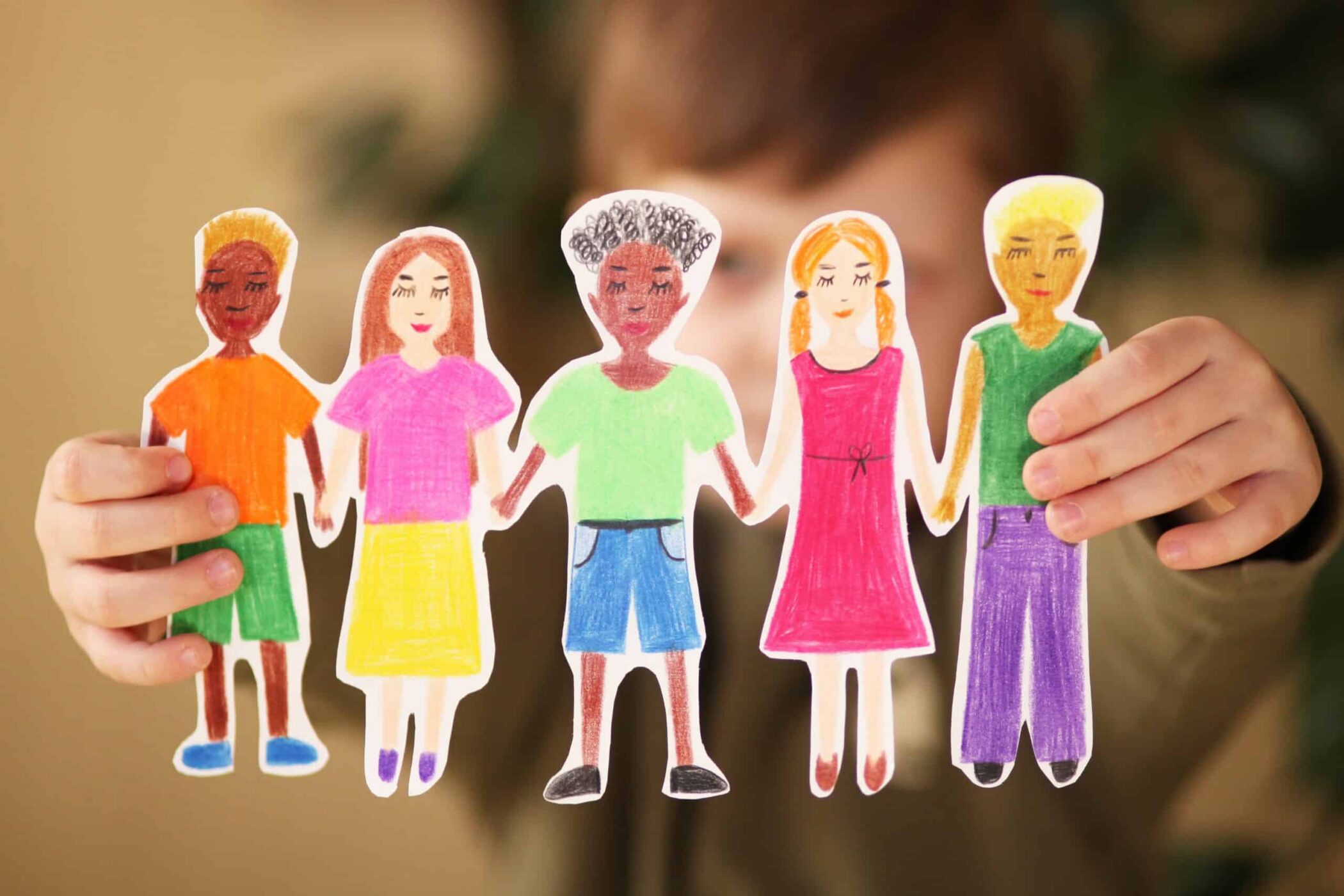 A child holds paper cut-outs of five diverse, hand-drawn people, smiling and holding hands, symbolising friendship and unity.