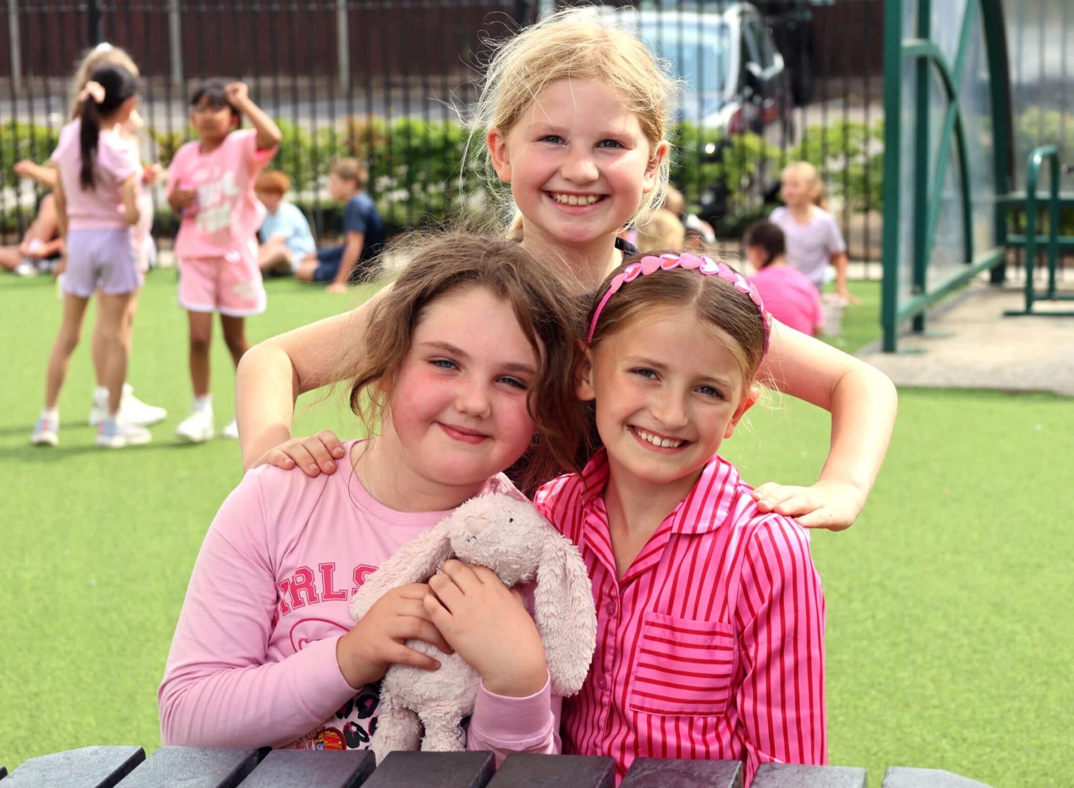 Three young girls smiling outdoors, two standing and one sitting holding a stuffed bunny. Other children play in the background on a grassy playground, all dressed in colourful clothes.