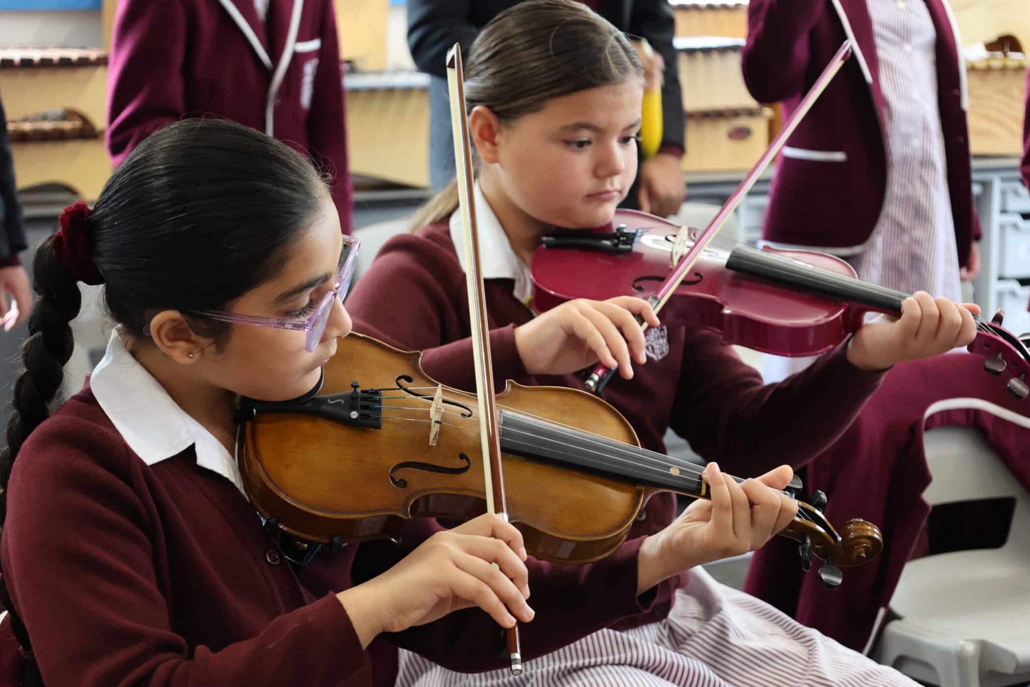 Two girls in maroon school uniforms play violins in a classroom setting, focused on their instruments. Other pupils are visible in the background, reflecting the lively atmosphere often seen at Open Events.