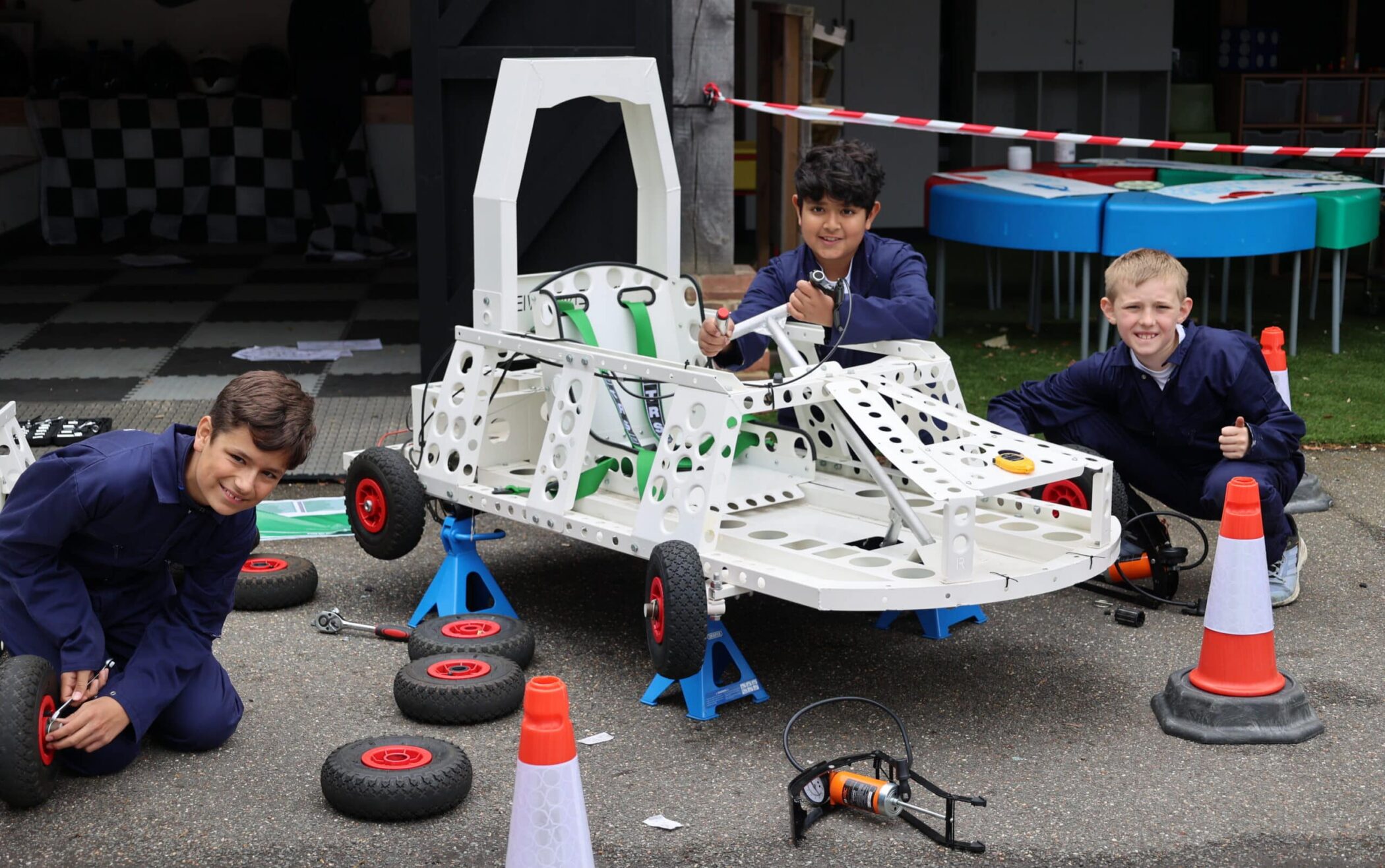 Three boys in blue overalls work together assembling a white go-kart with green seats at an Open Events gathering. Spare tyres and tools surround them outdoors, with traffic cones and a chequered flag backdrop visible.