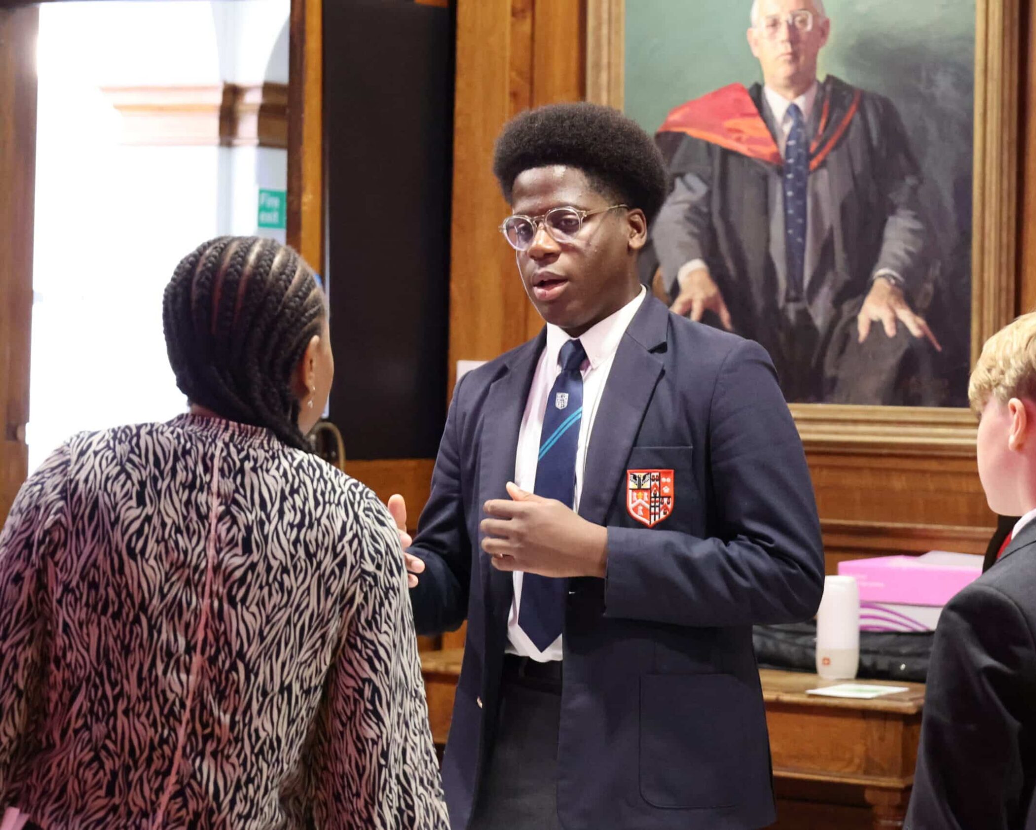 Three pupils in school uniforms converse indoors in front of a large framed portrait of a man in academic robes. One pupil speaks whilst others listen attentively, reflecting the formal, academic atmosphere found at Open Events.